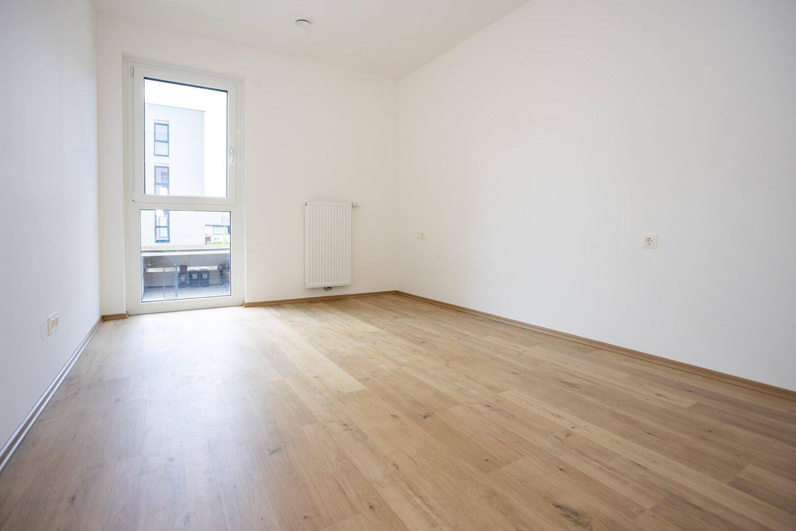 Leeres Zimmer mit hellem Laminatboden, weißen Wänden und Zugang zu einem Balkon.