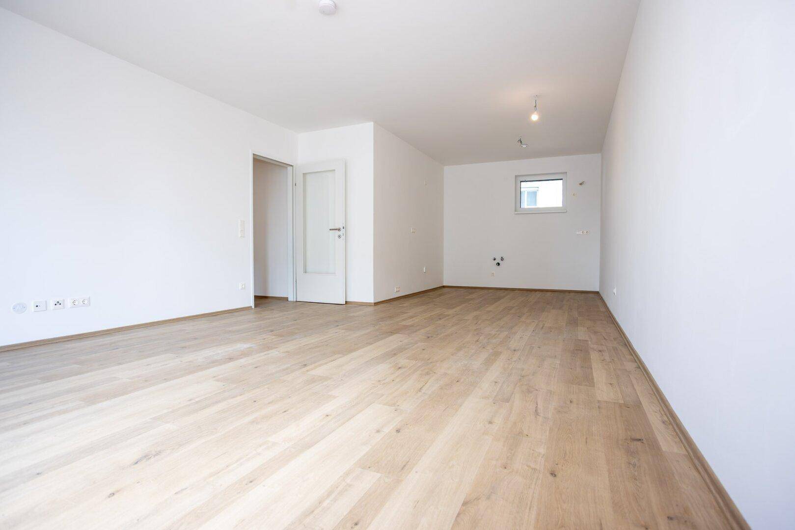 Geräumiges Wohnzimmer mit hellem Laminatboden, weißen Wänden und viel natürlichem Licht.