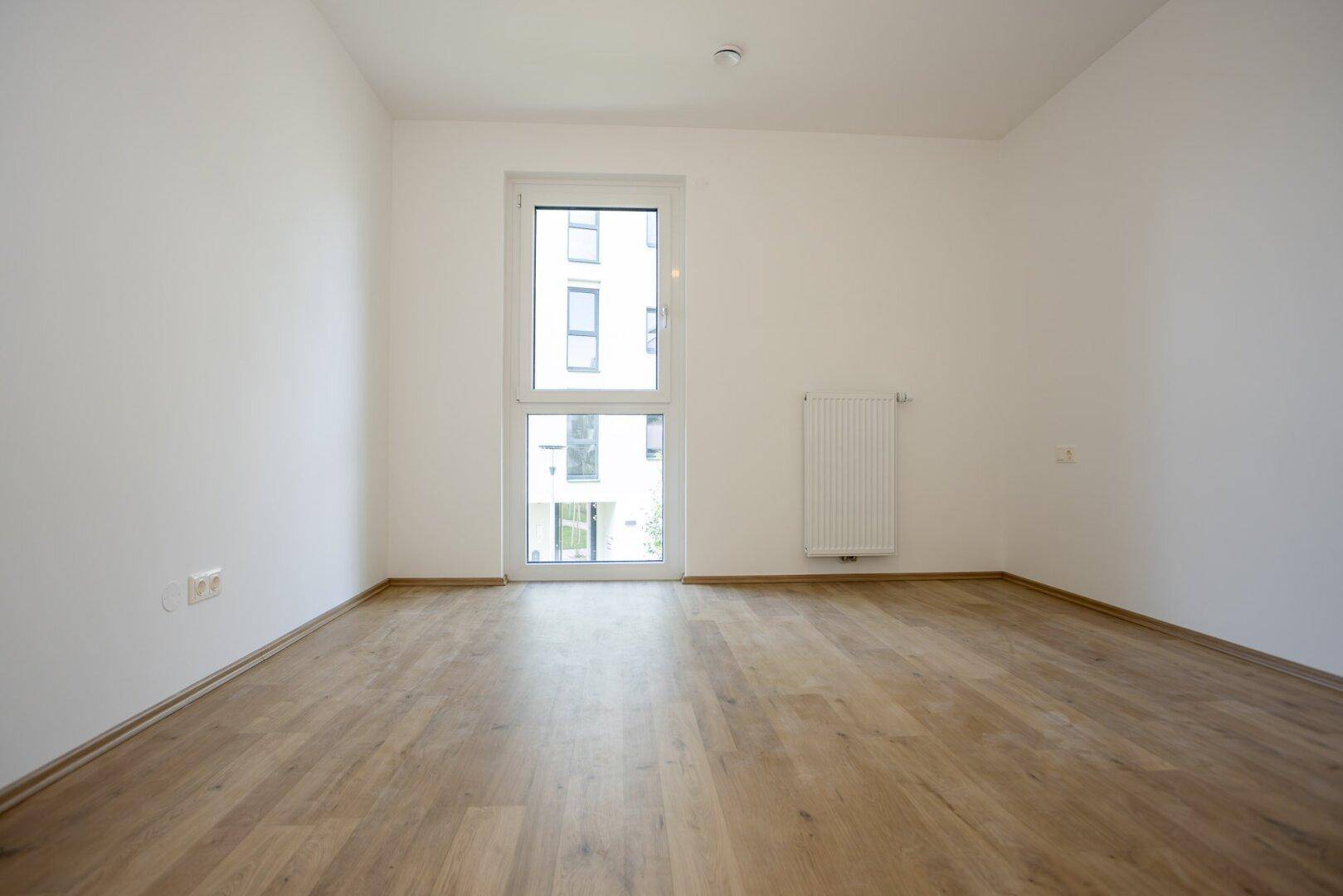 Helles, leeres Zimmer mit Laminatboden und großem Fenster, ideal als Schlafzimmer.