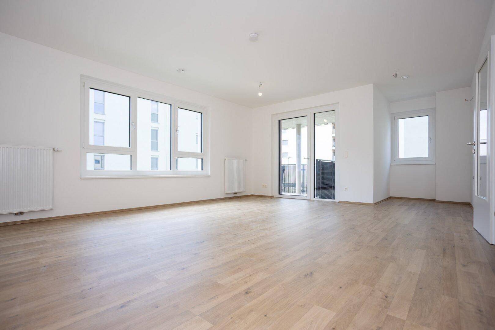 Großzügiges Wohnzimmer mit hellem Laminatboden, großen Fenstern und Balkonzugang.