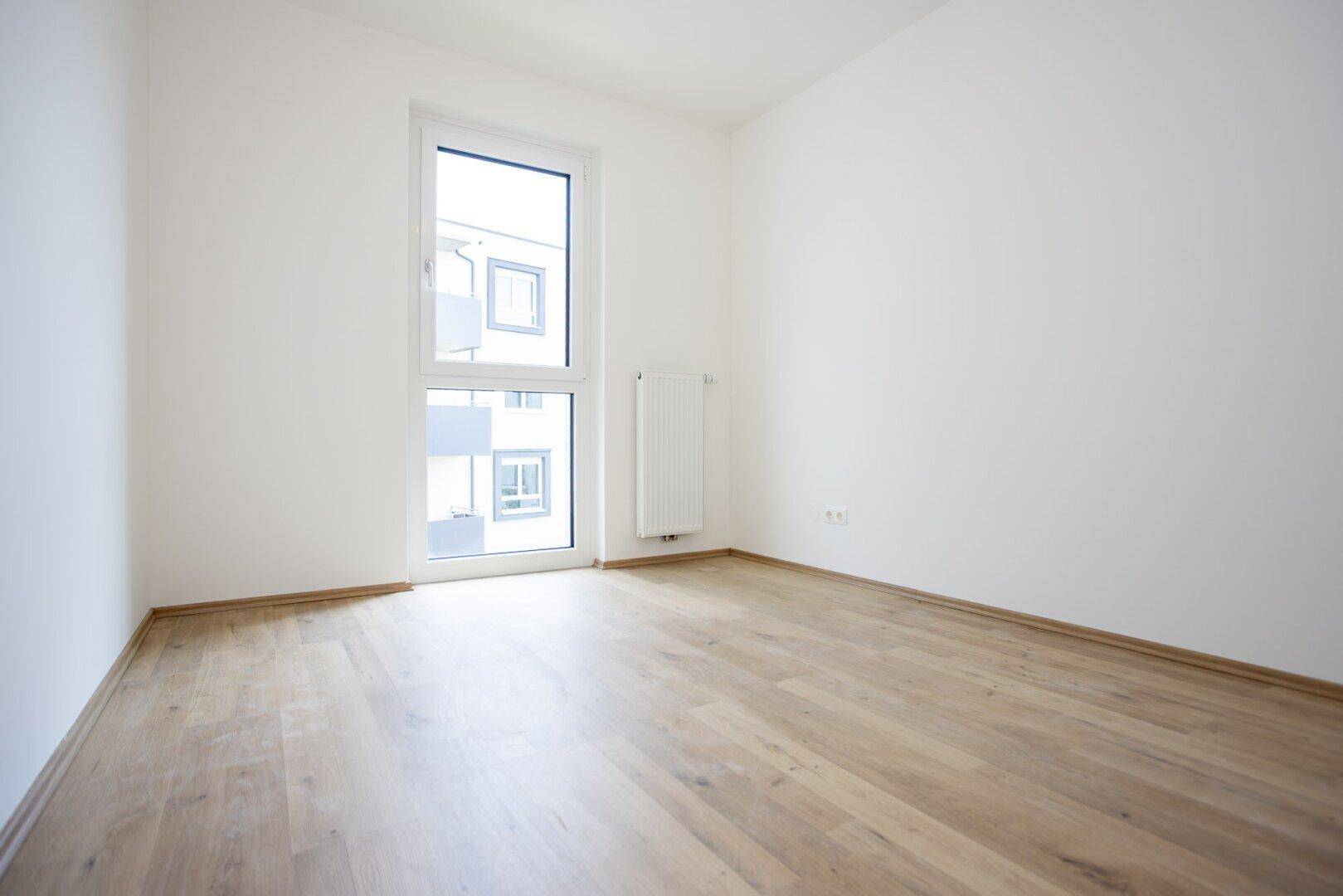 Leeres, helles Zimmer mit Laminatboden und einem hohen Fenster, das viel Tageslicht hereinlässt.