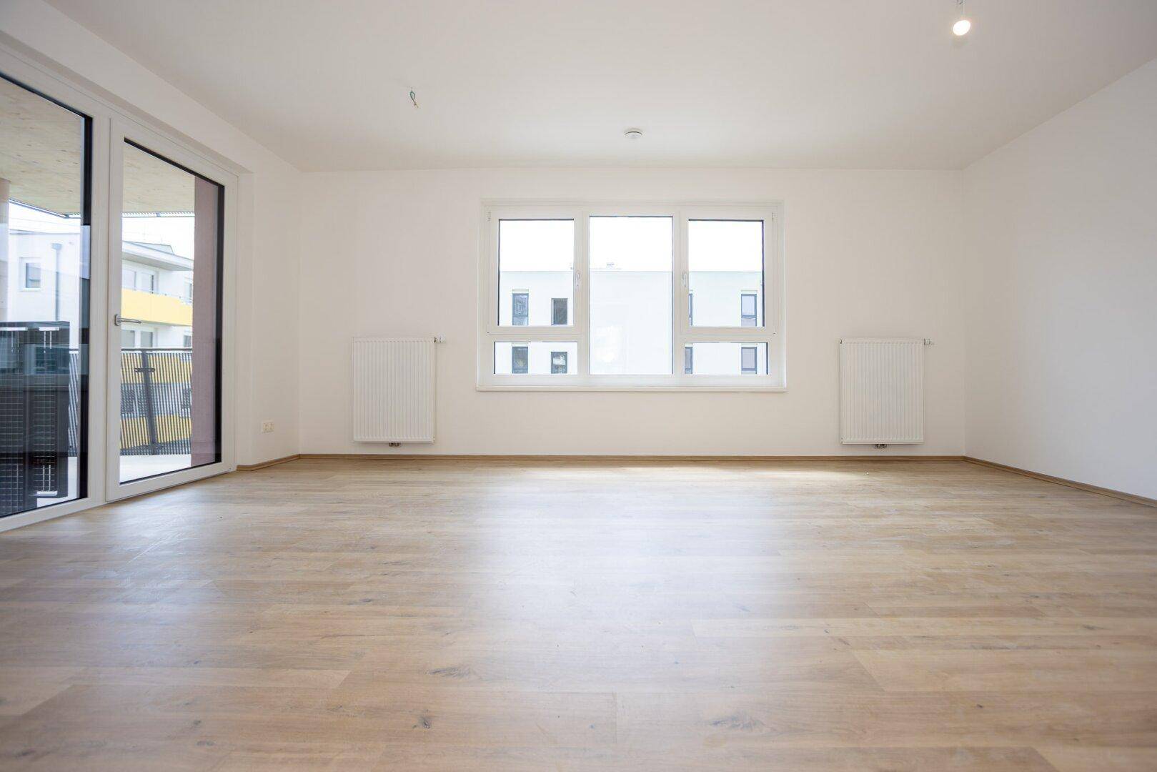 Geräumiger, heller Wohnbereich mit Laminatboden, großen Fenstern und direktem Zugang zu einem Balkon.