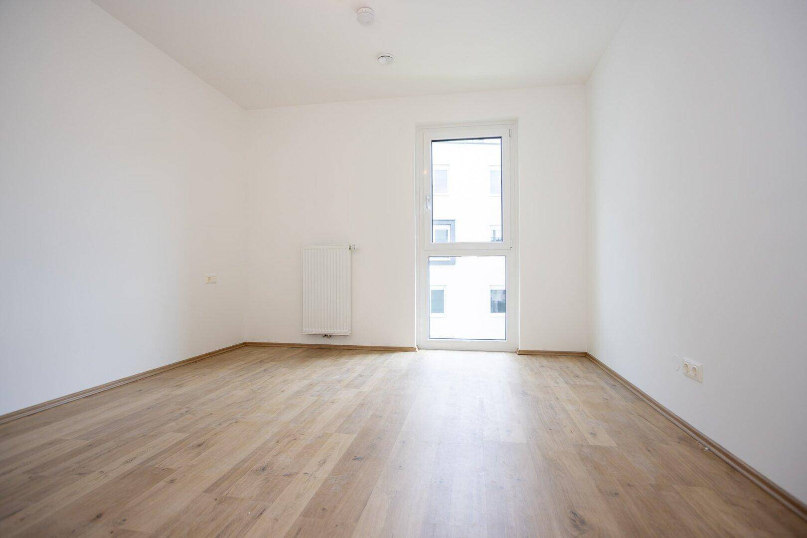 Leeres, helles Zimmer mit Laminatboden und einem hohen Fenster, das viel Licht in den Raum bringt.