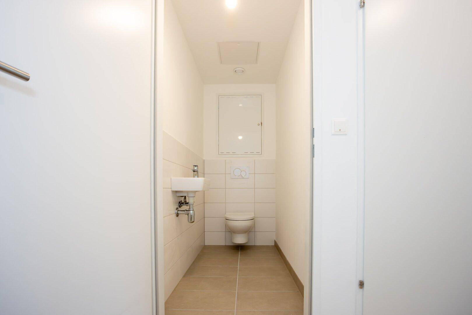 Kompaktes Gäste-WC mit Wand-WC, kleinem Waschbecken und hellen Fliesen in modernem Design.