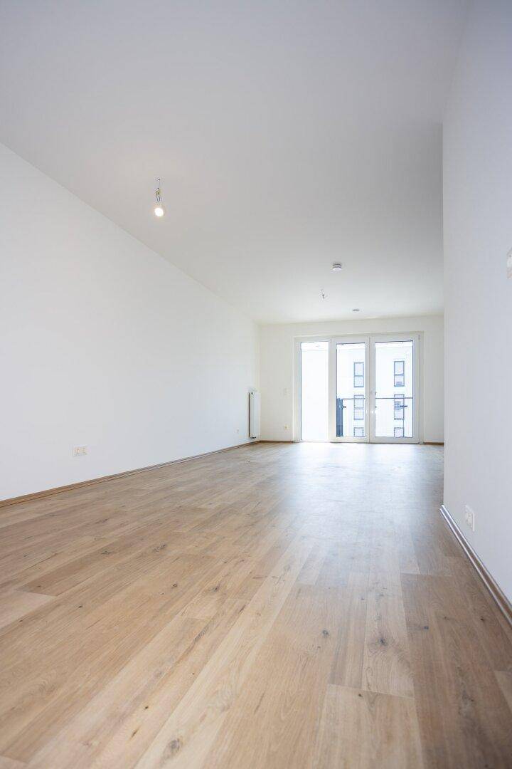 Geräumiges, leeres Wohnzimmer mit hellem Holzboden und großen Fenstern mit Balkonzugang.