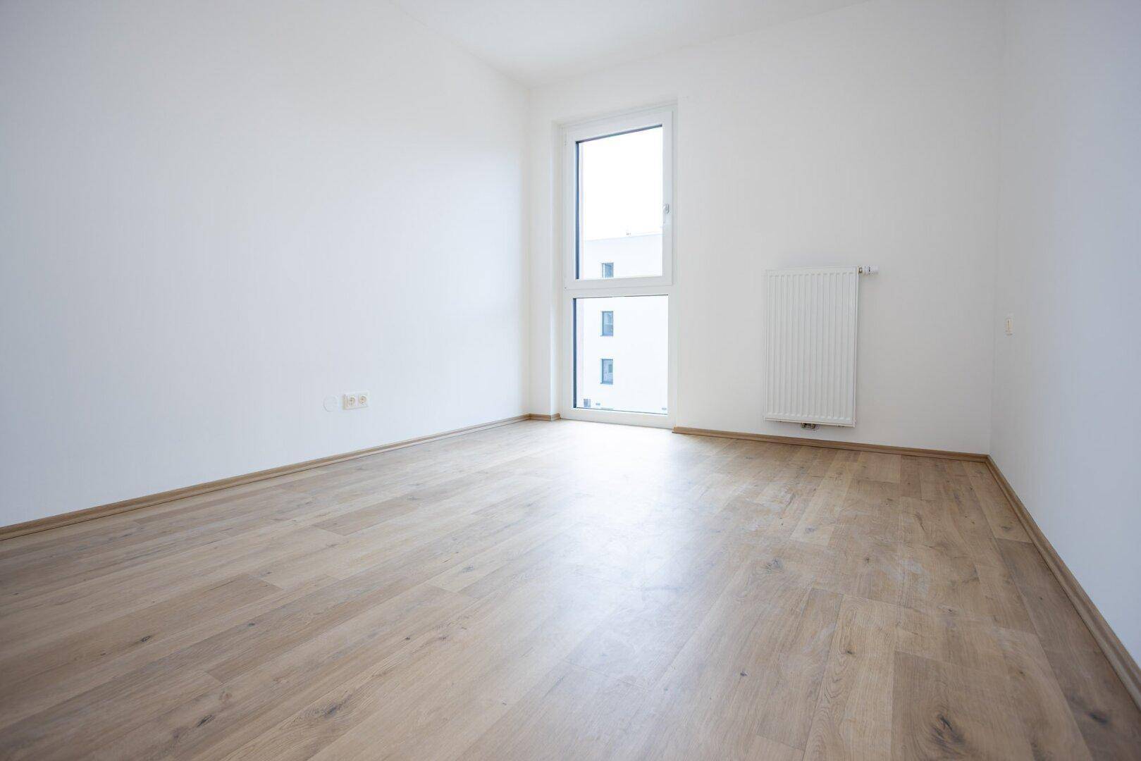 Leeres Schlafzimmer mit hellem Holzboden, großem Fenster und Heizkörper, bereit zum Einzug.