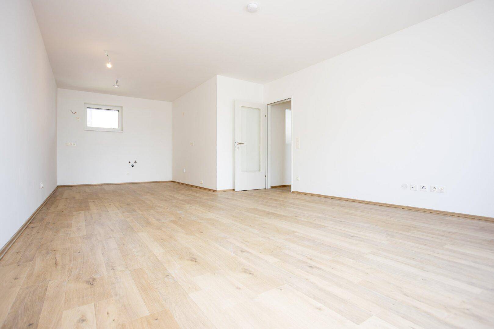 Leerer Raum mit Laminatboden, weiß gestrichenen Wänden und einem kleinen Fenster.