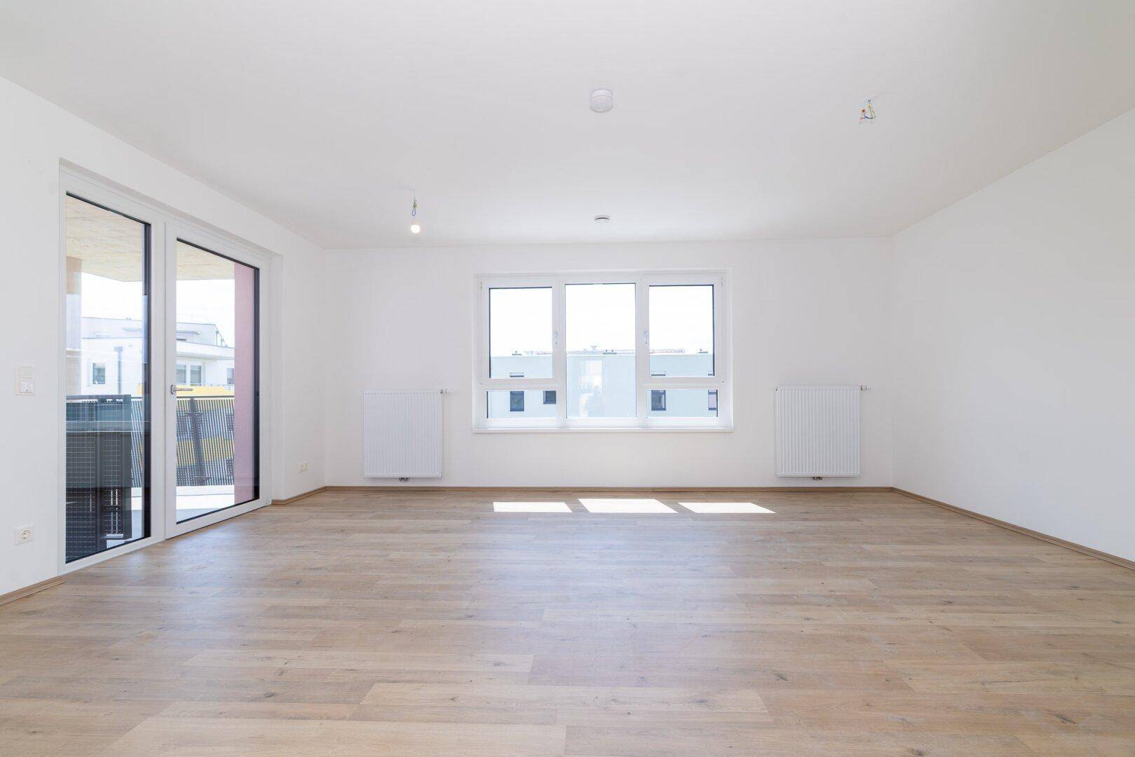 Lichtdurchfluteter Wohnbereich mit großen Fenstern, Zugang zum Balkon und hellem Laminatboden.