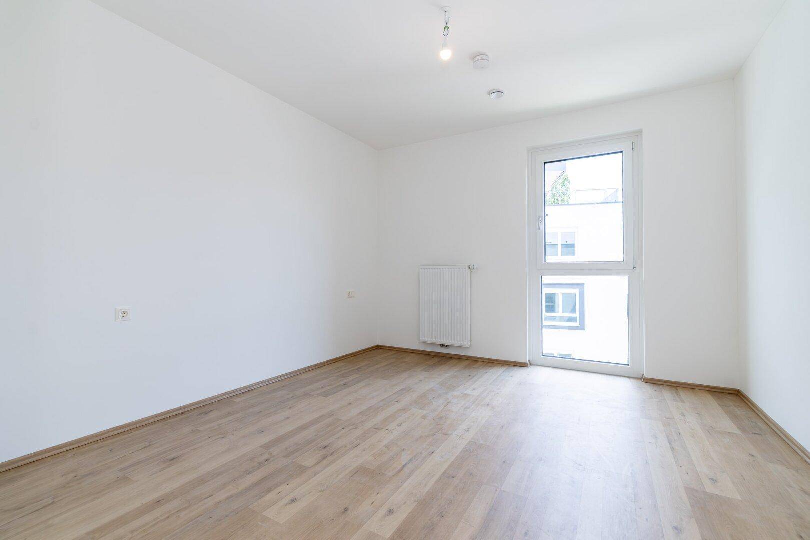 Helles Schlafzimmer mit Laminatboden und einem großen Fenster, das viel Tageslicht hereinlässt.