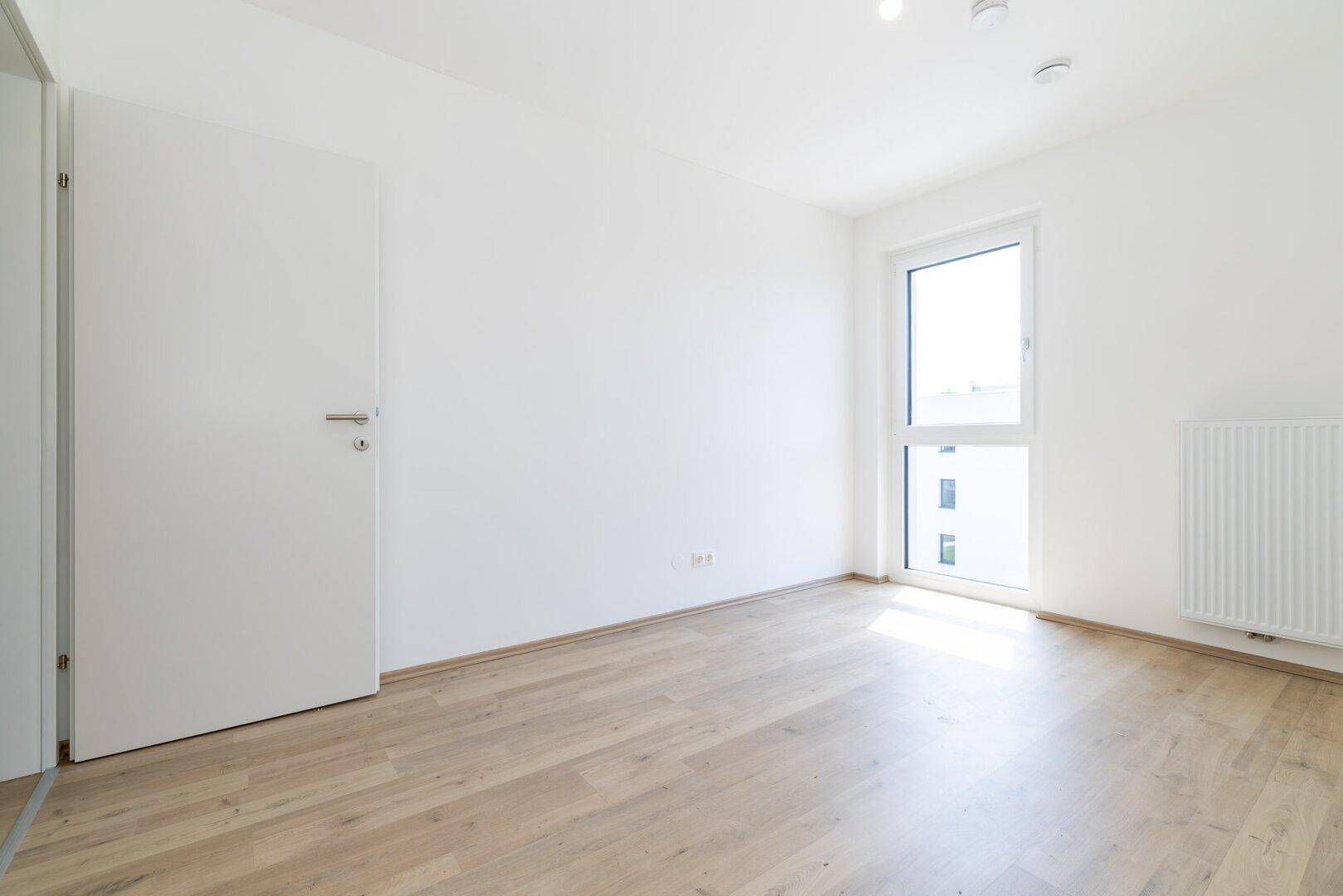 Helles Schlafzimmer mit Laminatboden und großem Fenster, das viel Tageslicht hereinlässt.