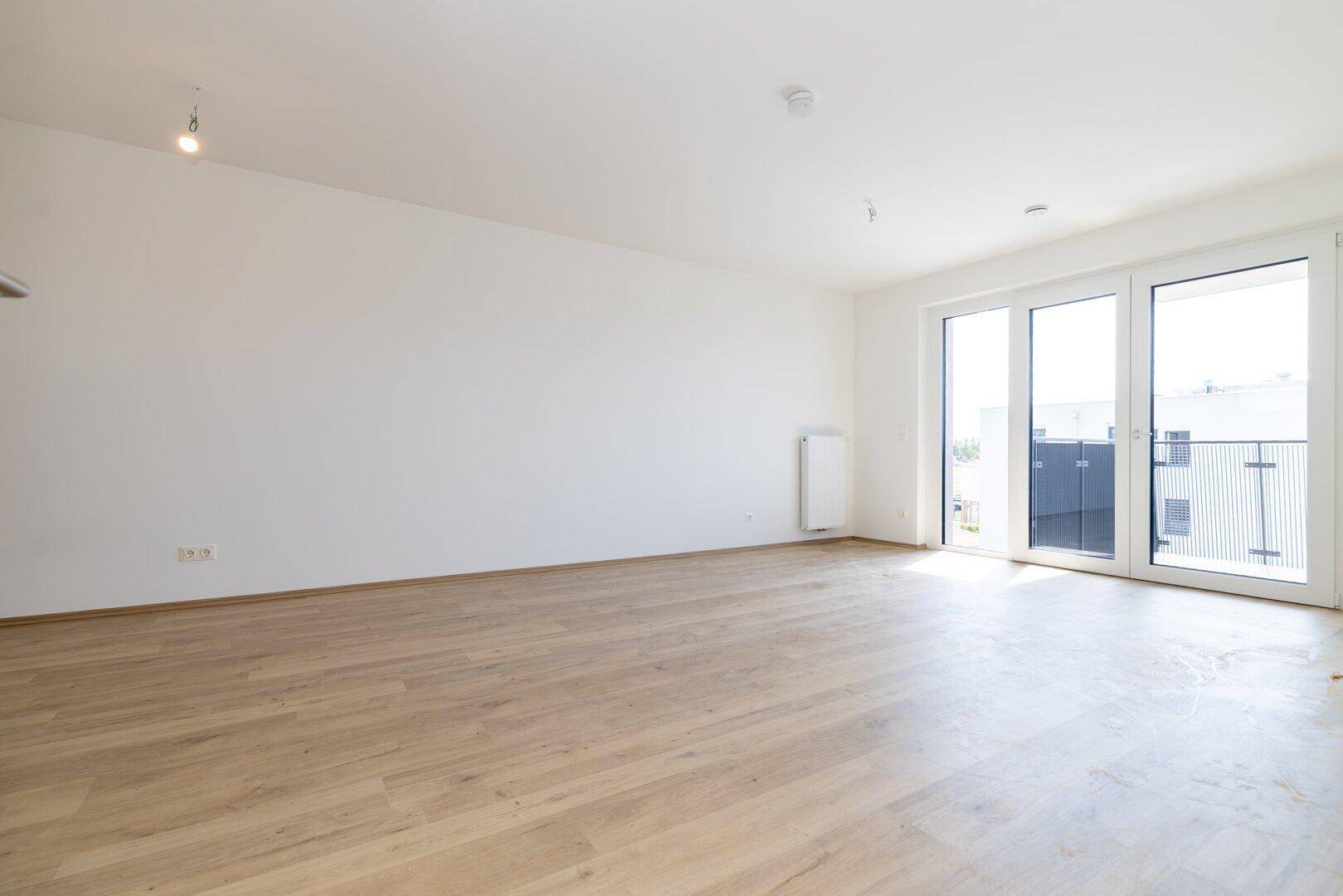 Geräumiges Wohnzimmer mit Laminatboden und großen Glastüren, die zu einem Balkon führen.