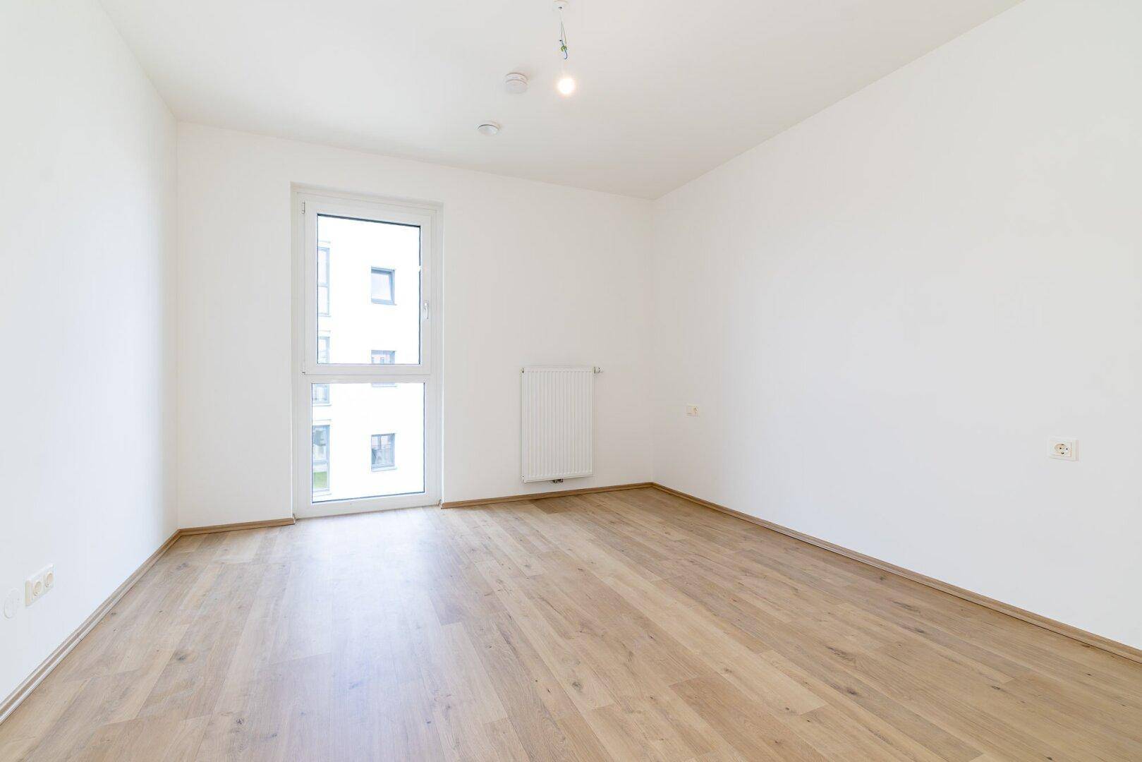 Heller, leerer Raum mit Laminatboden und einem großen Fenster, das viel Licht hereinlässt.
