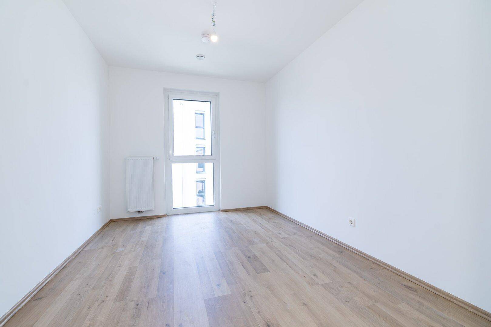Leerer Raum mit hellem Laminatboden und einem Fenster, das den Blick nach draußen freigibt.