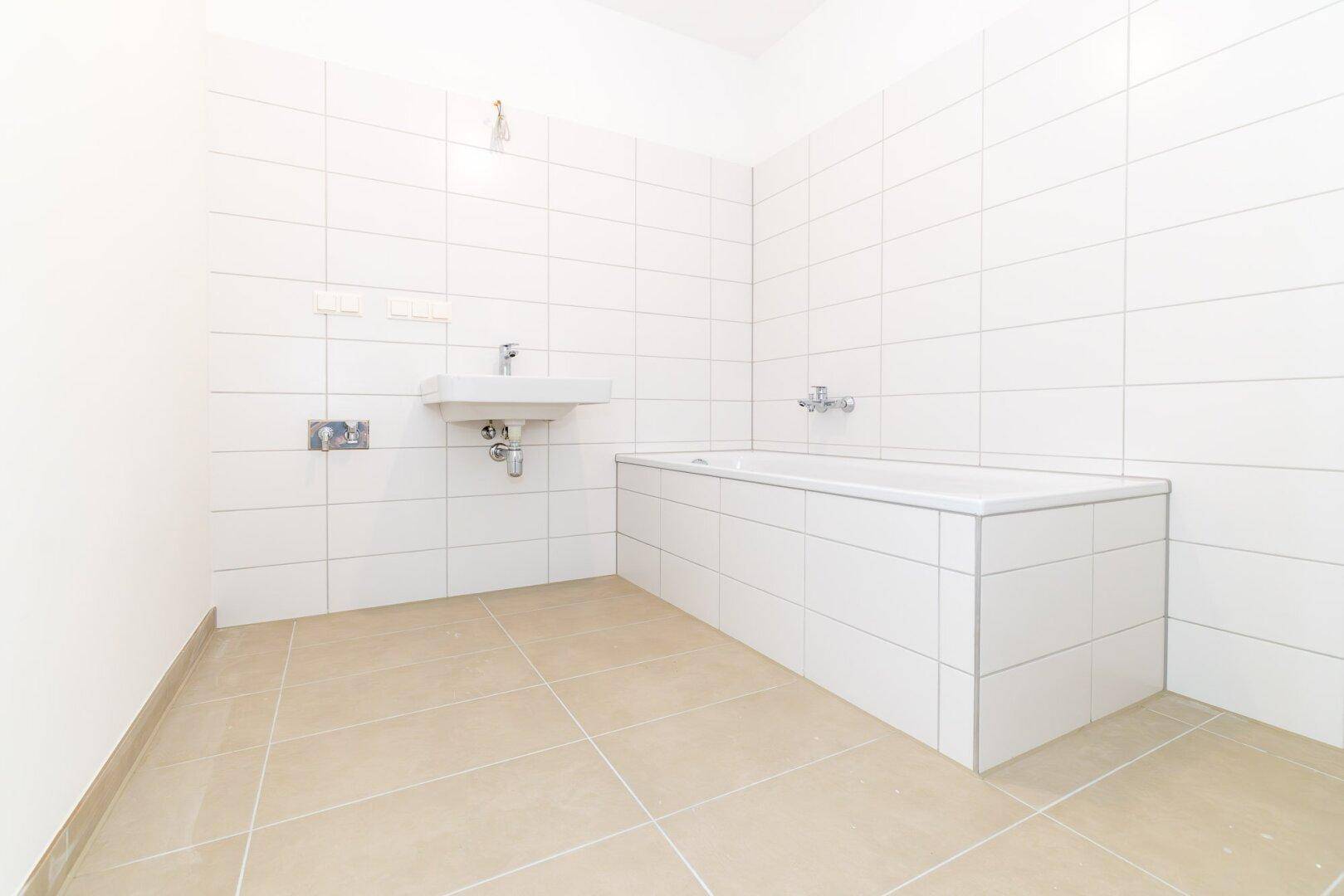 Helles Badezimmer mit Badewanne, Waschbecken und neutralen Fliesen an Wänden und Boden.