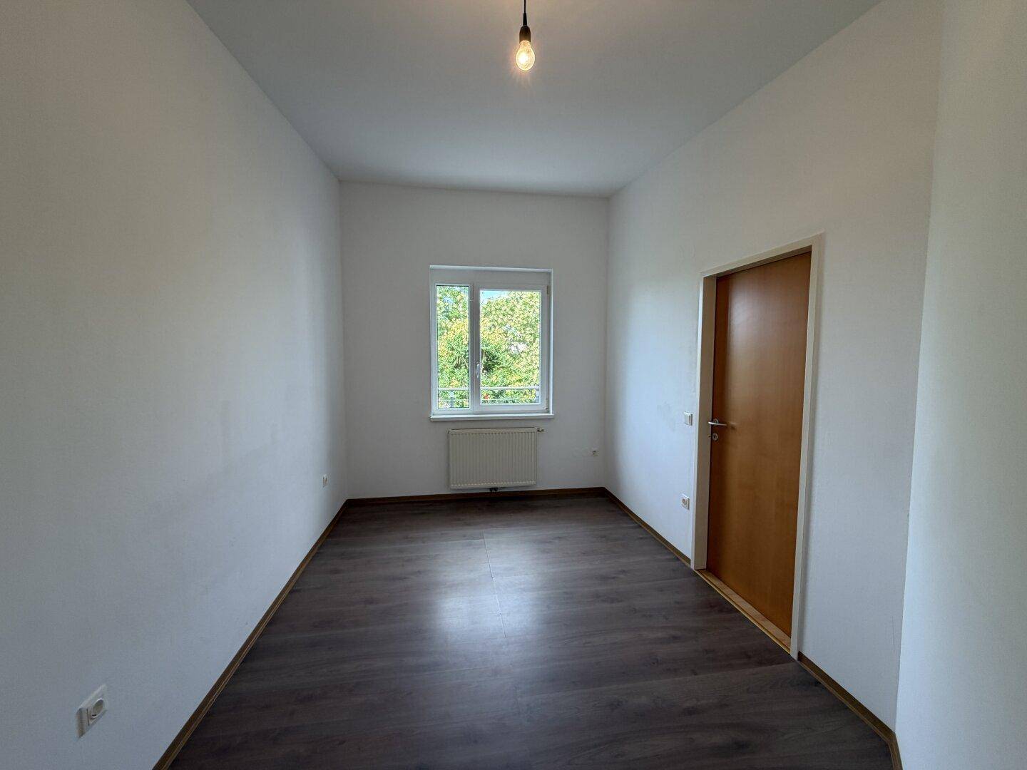 Helles Schlafzimmer mit dunklem Laminatboden und Fenster mit Blick ins Grüne.