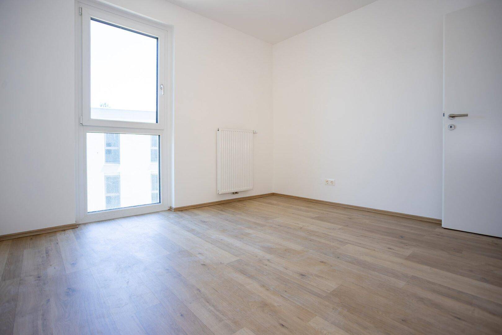 Leeres Schlafzimmer mit hellem Laminatboden und großem Fenster.