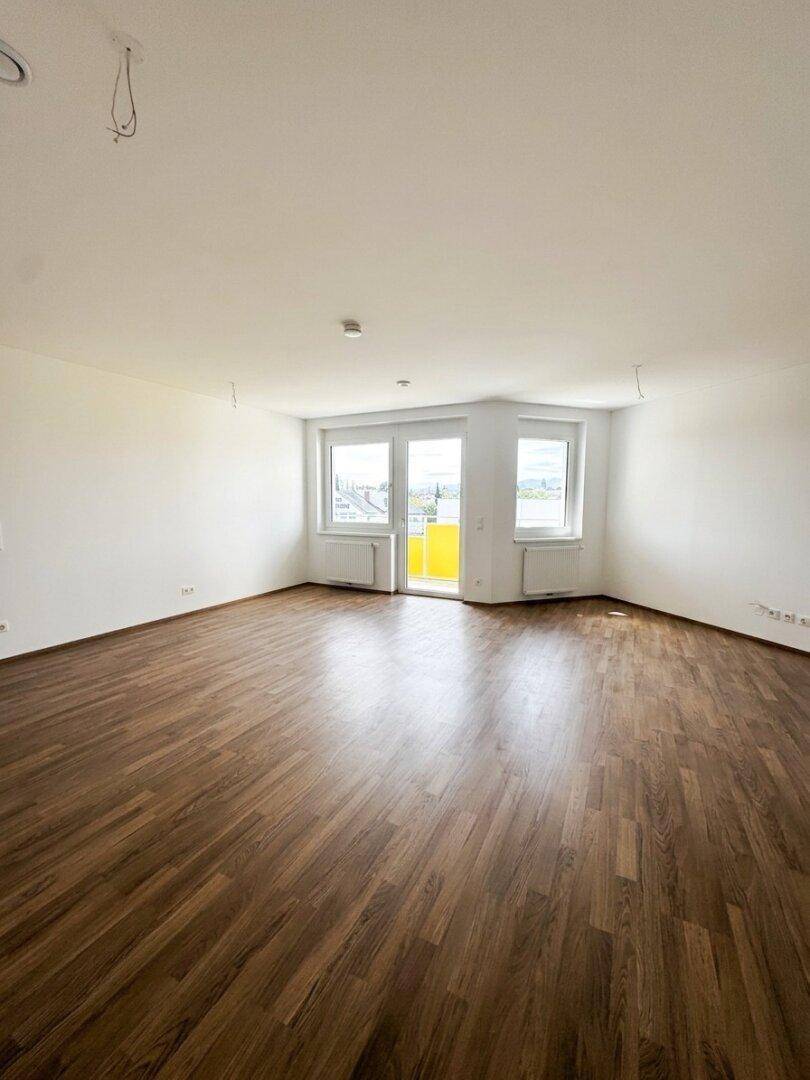 Heller, leerer Wohnraum mit modernem Laminatboden, weißen Wänden und Zugang zu einem Balkon.
