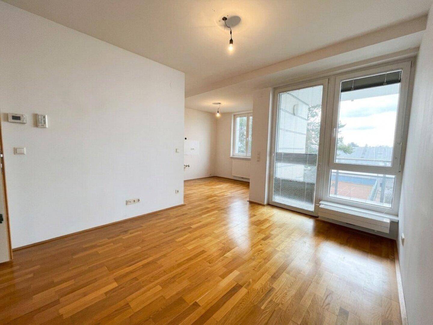 Geräumiger, heller Wohnraum mit glänzendem Parkettboden und großen Fenstern, die viel Tageslicht hereinlassen.