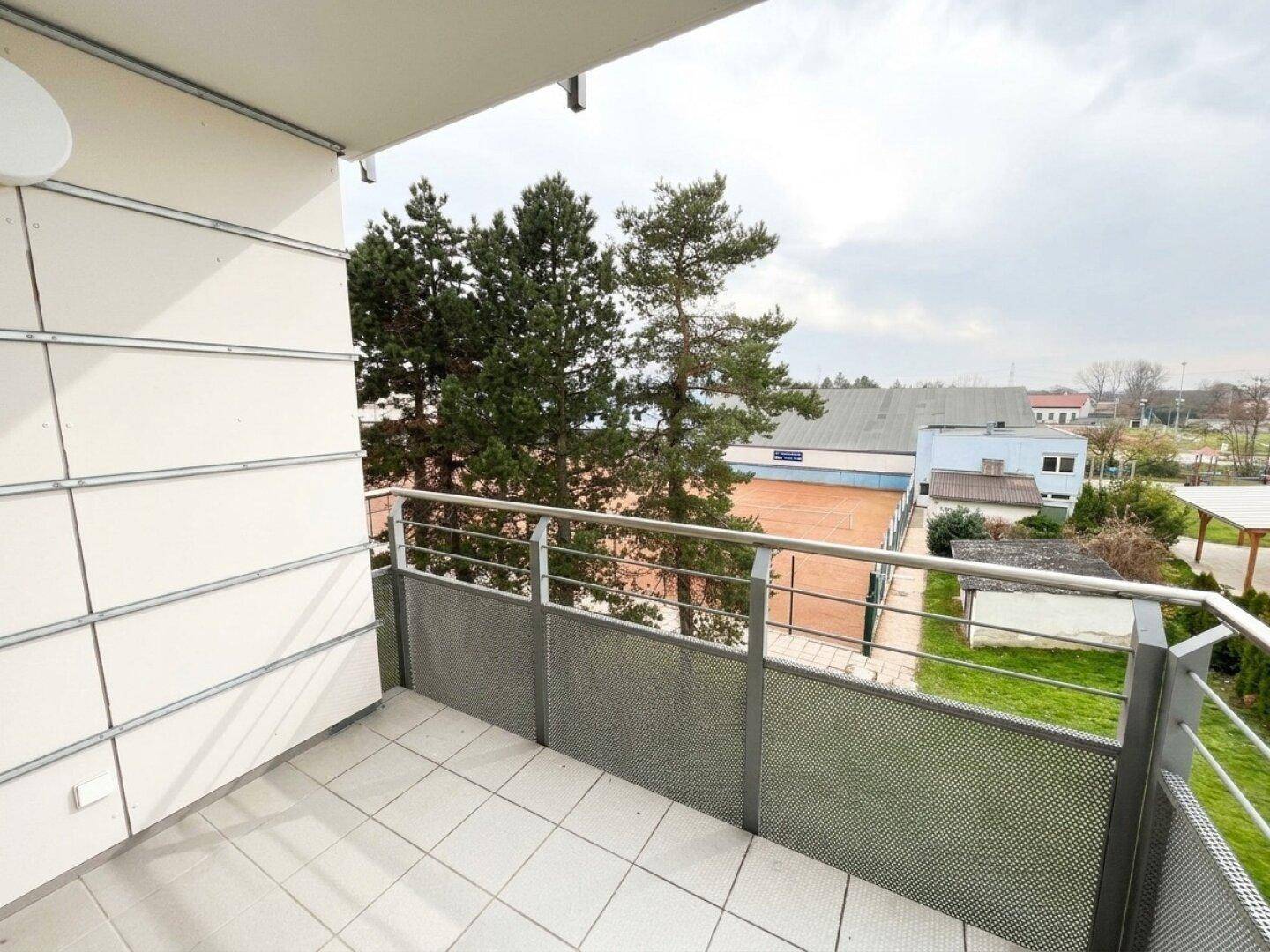Balkon mit Fliesenboden und Blick auf grüne Bäume und einen nahegelegenen Tennisplatz.