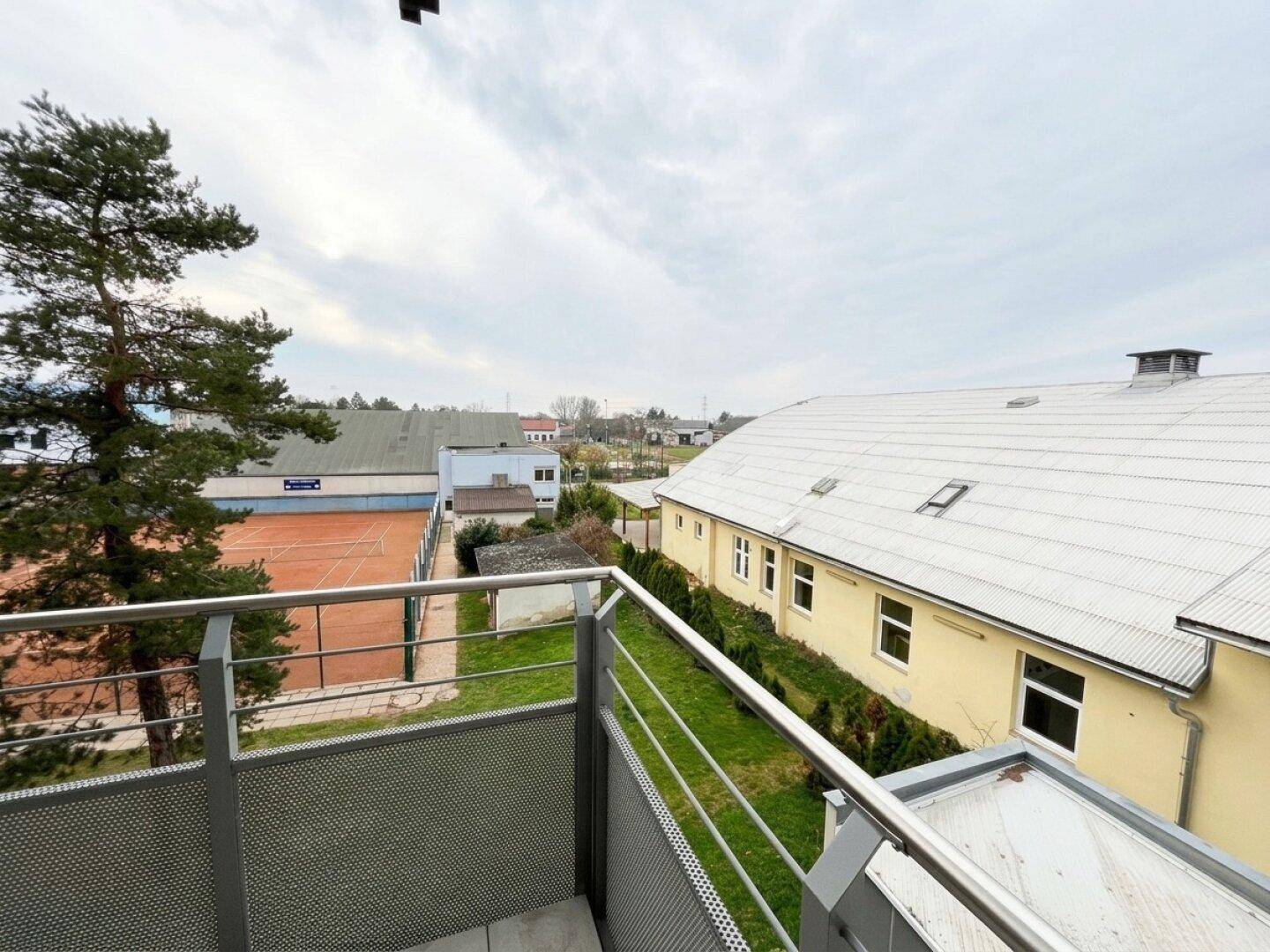 Balkon mit Fliesenboden und weitem Blick auf Bäume, einen Tennisplatz und umliegende Gebäude.