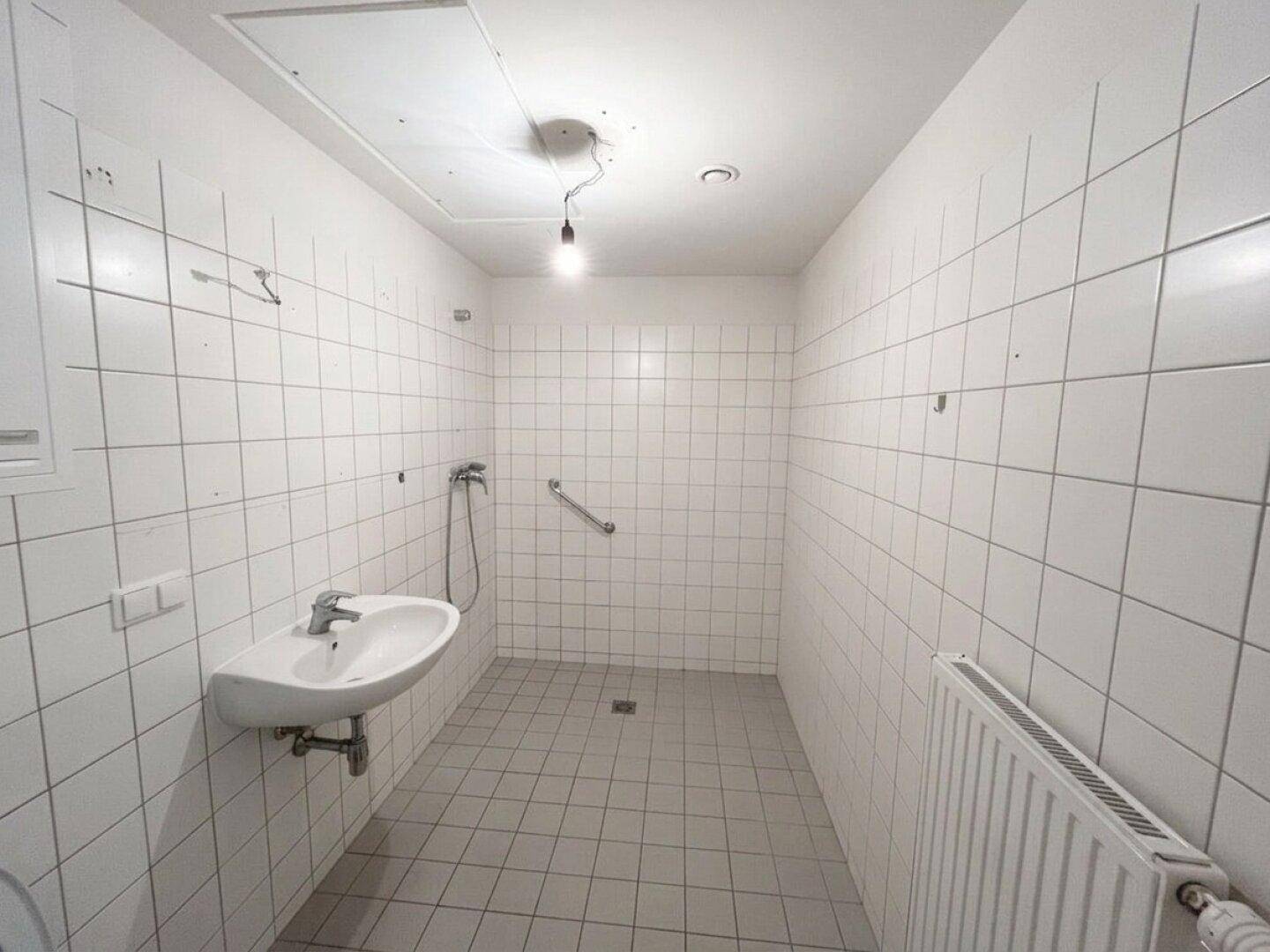 Funktionales Badezimmer mit weißen Fliesen, Waschbecken, Heizkörper und Haltegriff in der Duschzone.