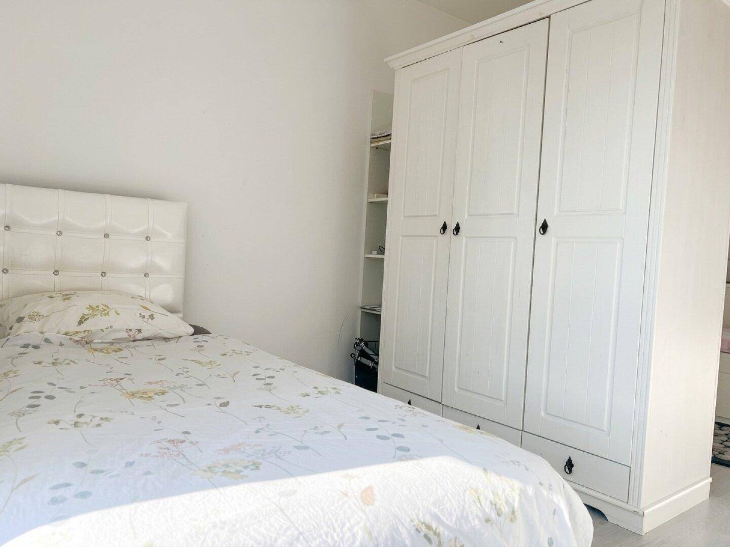 Helles Schlafzimmer mit weißem Bett, gepolstertem Kopfteil und passendem Kleiderschrank.