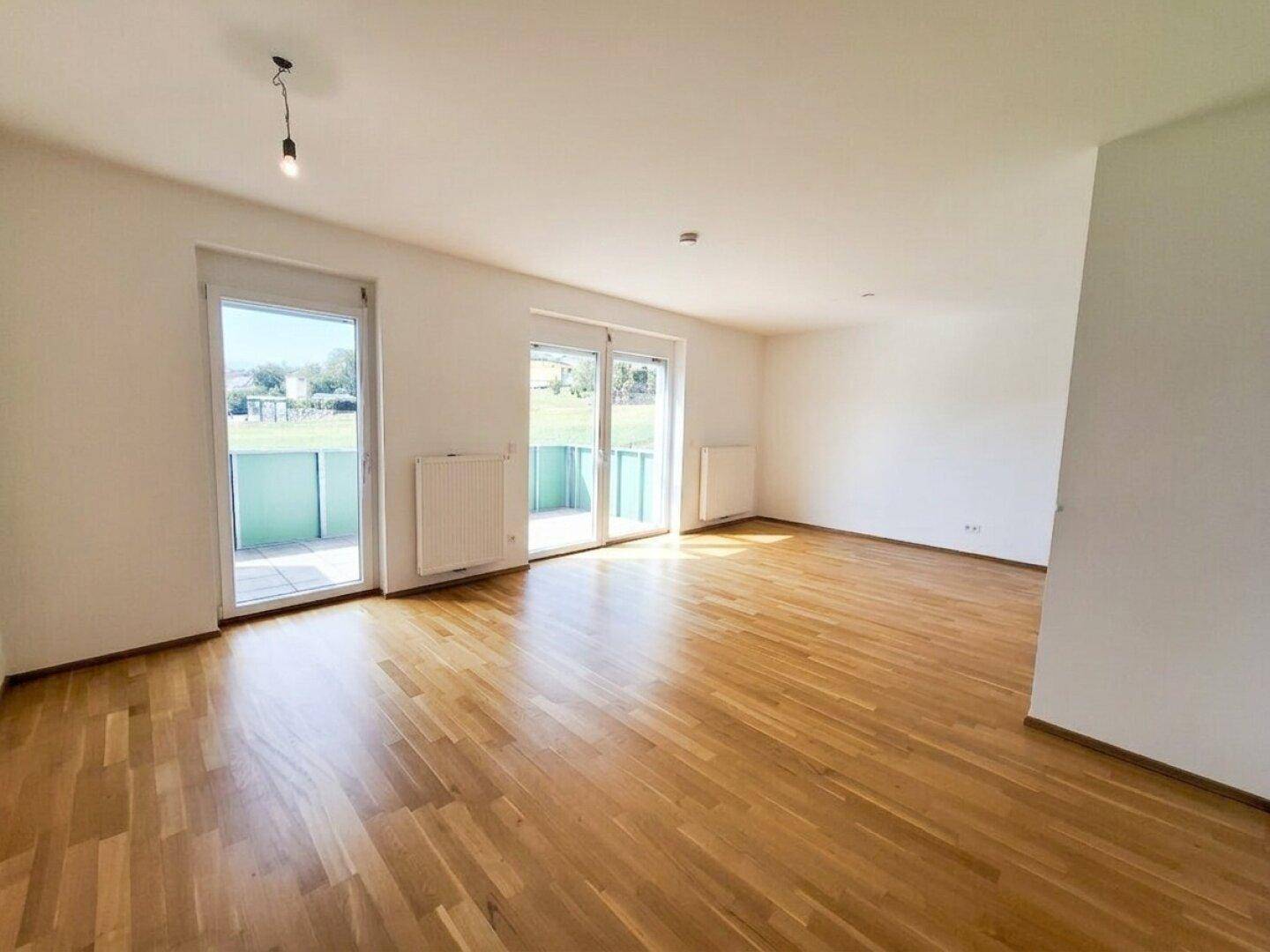 Helles, leeres Wohnzimmer mit warmem Parkettboden und zwei großen Fenstertüren zum Balkon.
