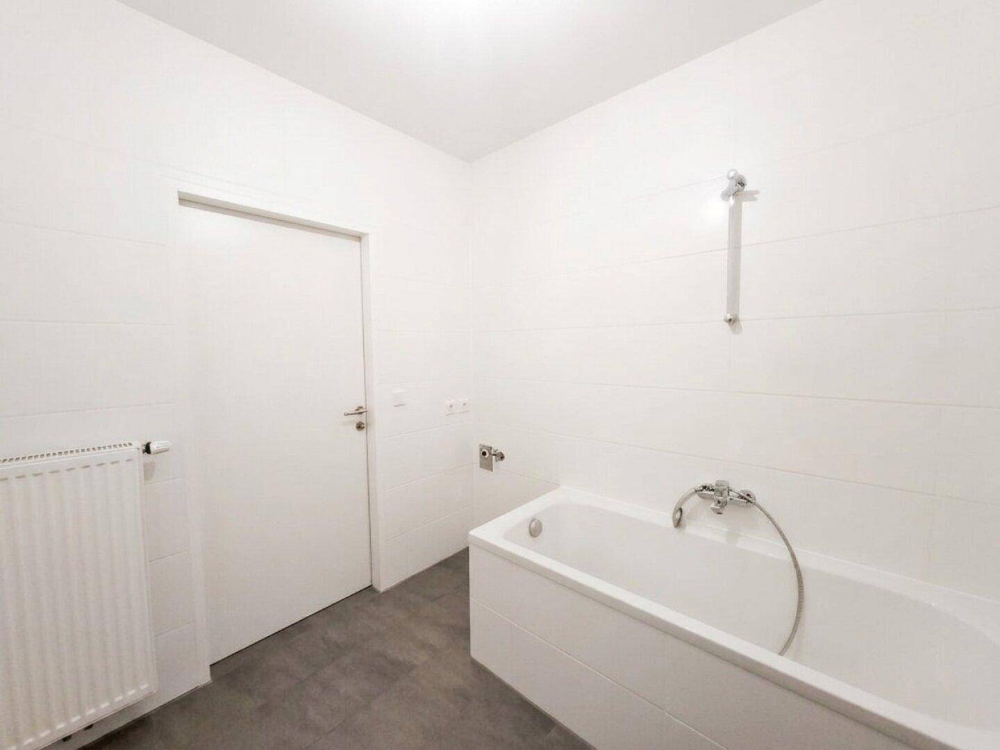 Helles Badezimmer mit Badewanne, Heizkörper und weiß gefliesten Wänden, bietet eine funktionale Ausstattung.