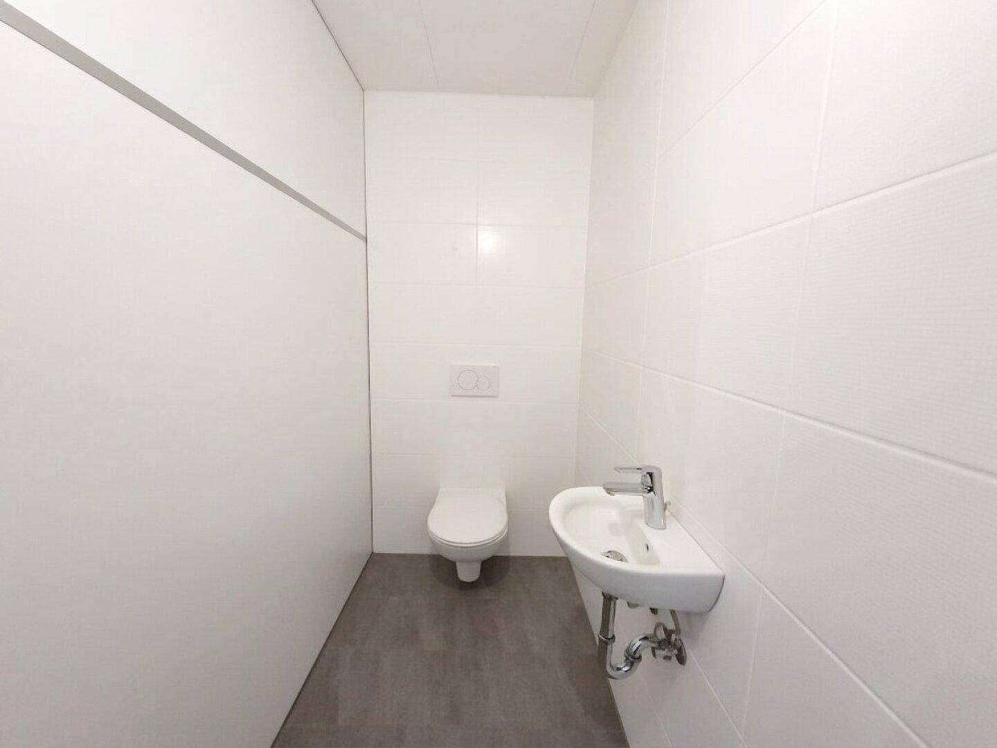 Separates WC mit wandhängender Toilette und kleinem Waschbecken, ausgestattet mit weißen Fliesen.
