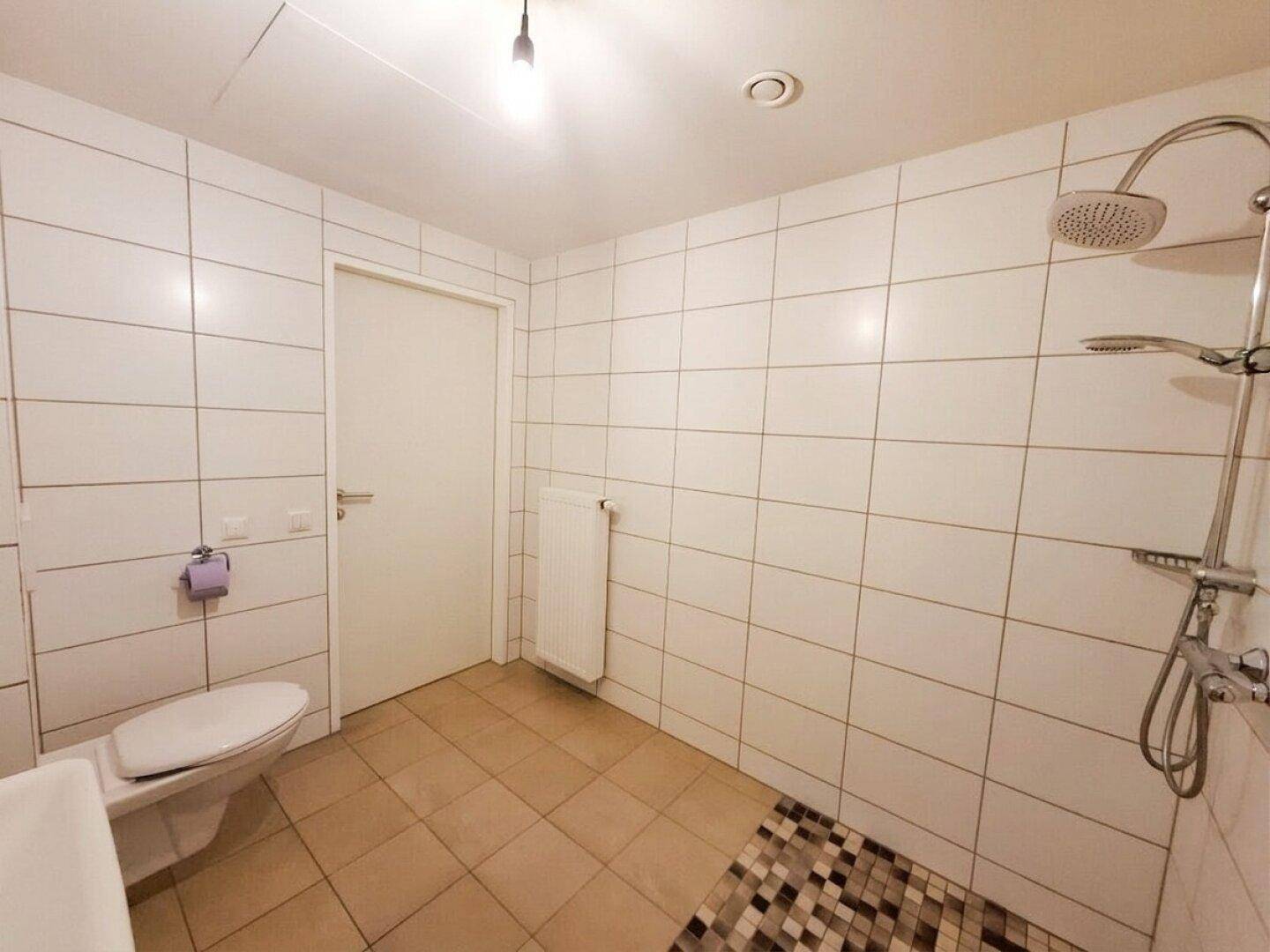 Funktionales Badezimmer mit Duschbereich, WC und Heizkörper, komplett gefliest in Weiß.