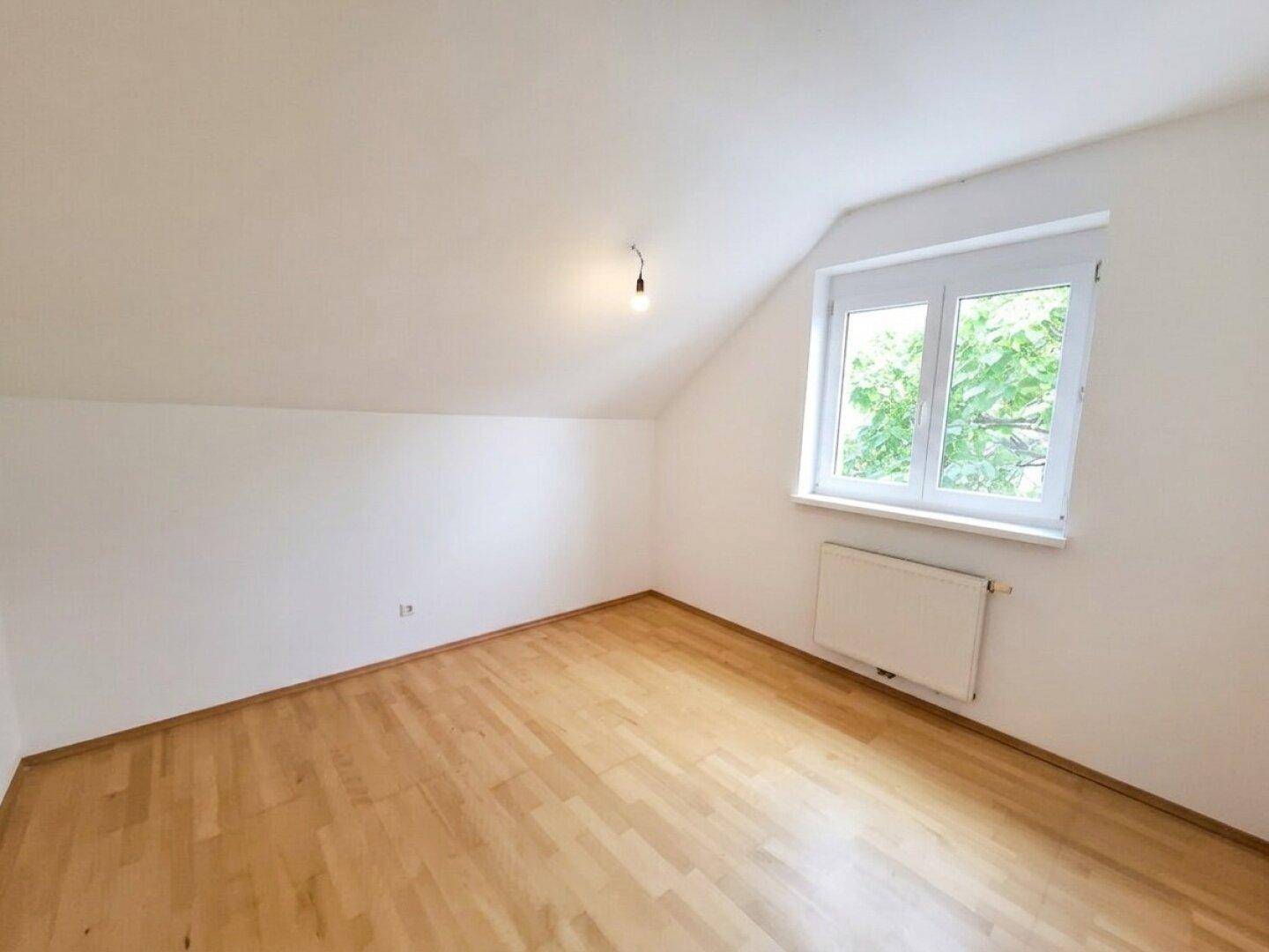 Dachzimmer mit schrägen Wänden, Parkettboden und einem Fenster mit Blick ins Grüne.