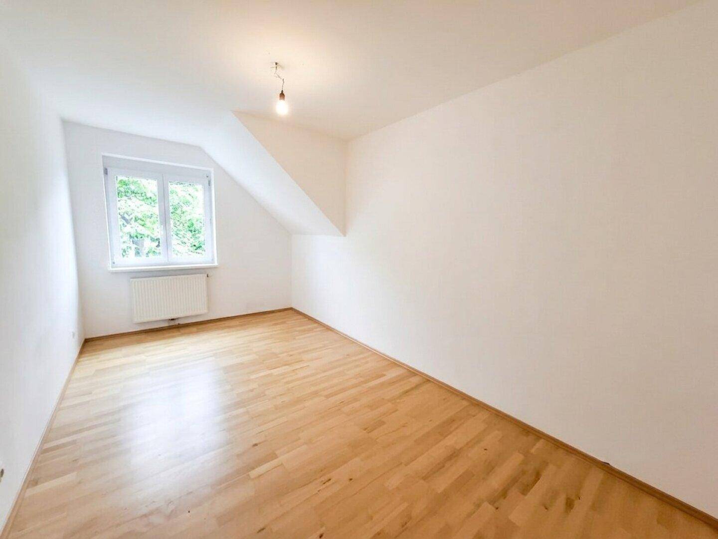 Helles Dachzimmer mit Parkettboden, schrägen Wänden und einem Fenster.