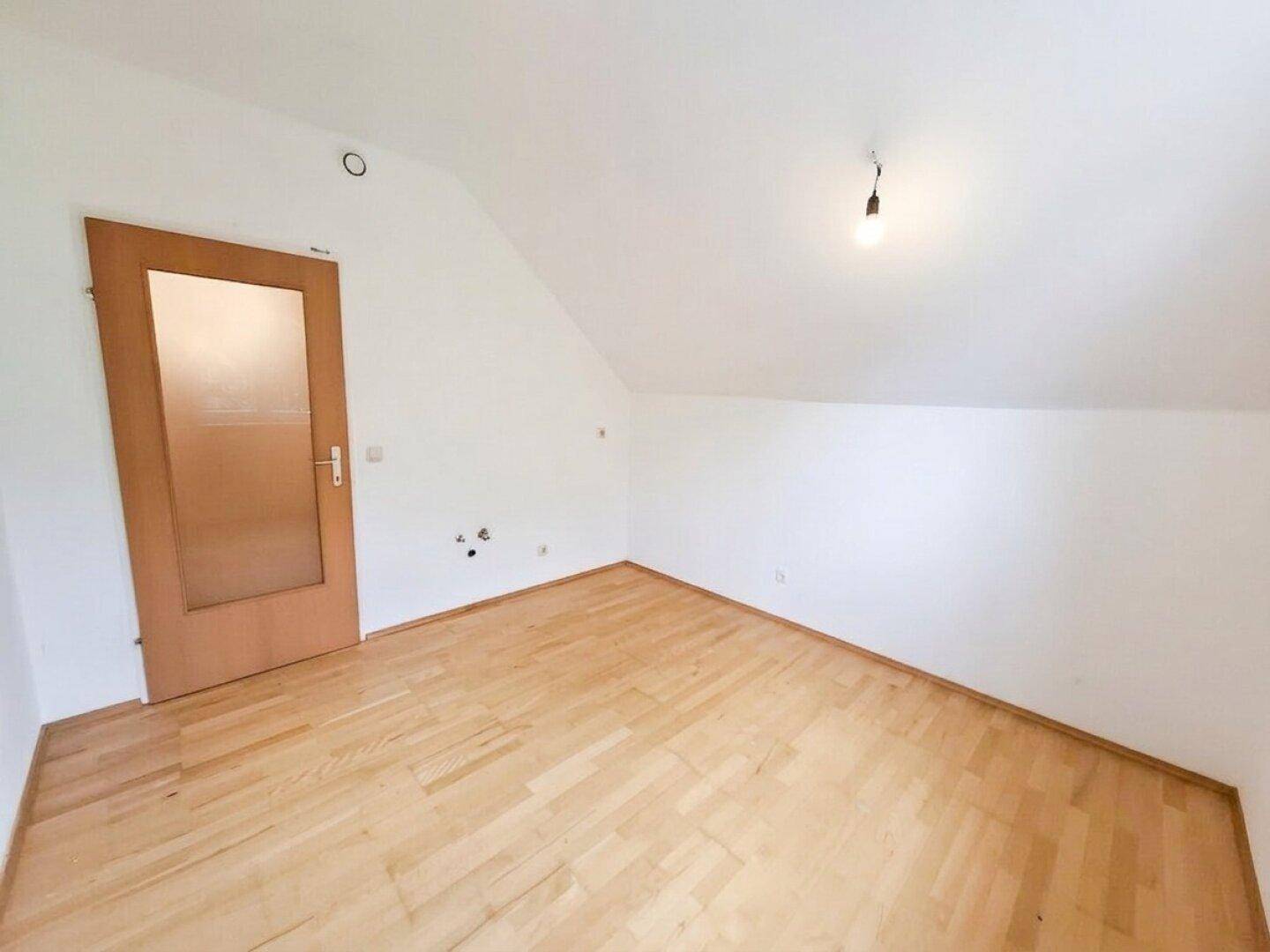 Helles Dachzimmer mit Parkettboden, schrägen Wänden und einer Holztür.