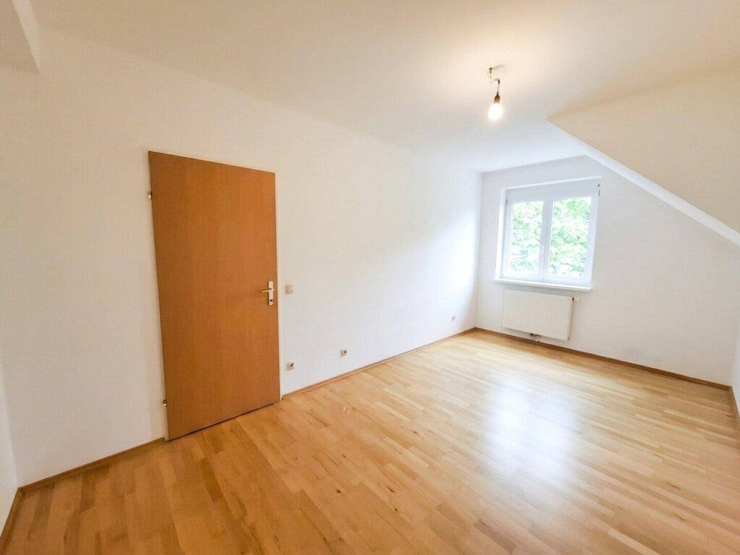 Dachzimmer mit Parkettboden, weißen Wänden, Holztür und Fenster.