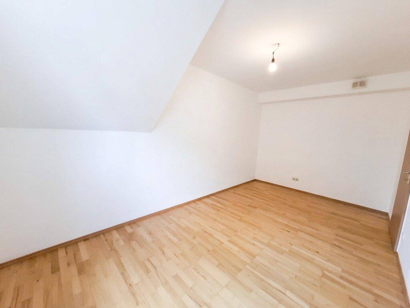 Geräumiges Dachzimmer mit Parkettboden, schrägen Wänden und einer Holztür.