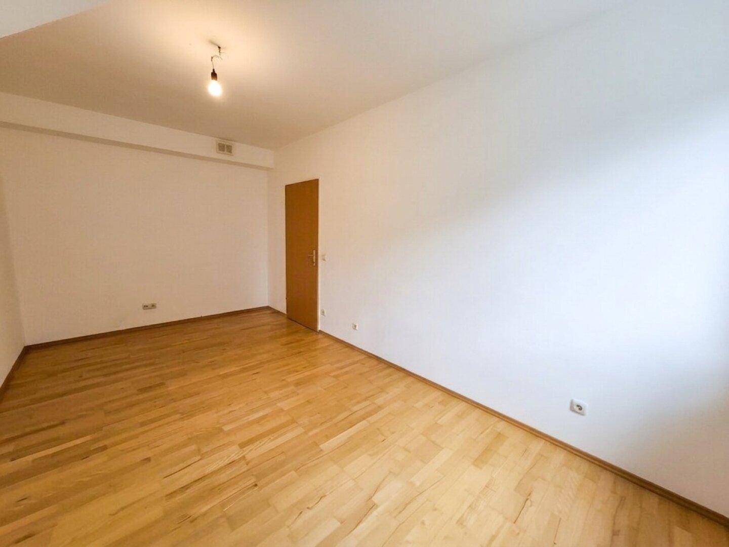 Helles Zimmer mit Parkettboden, weißen Wänden und einer schlichten Holztür.