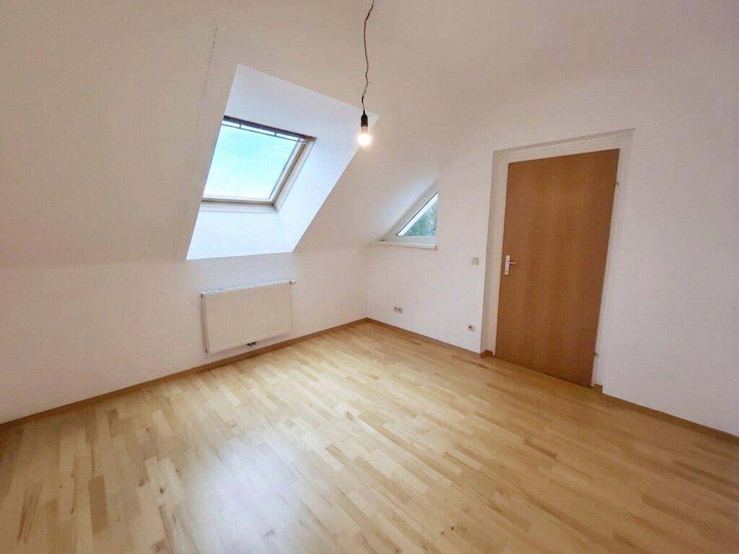 Helles Dachzimmer mit Parkettboden, Dachfenster und einer Holztür.