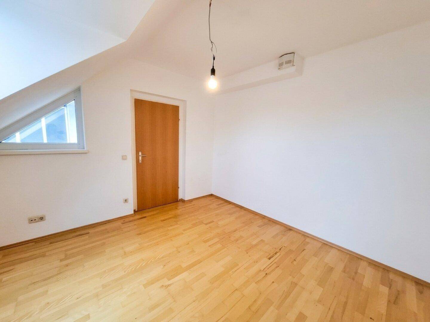 Dachzimmer mit Parkettboden, schrägen Wänden, Holztür und kleinem Fenster.