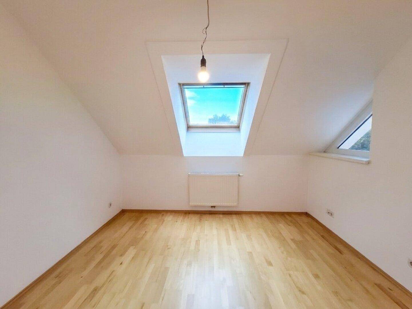 Helles Dachzimmer mit Parkettboden, schrägen Wänden und einem großen Dachfenster.