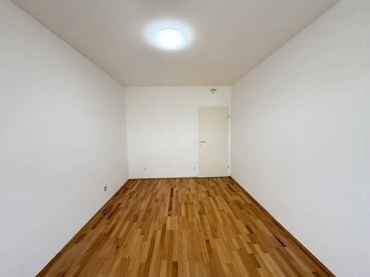 Schlichtes, leeres Zimmer mit Parkettboden und weiß gestrichenen Wänden.