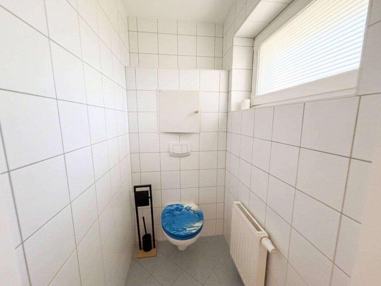 Separates WC mit Fenster, Wand-WC und weißen Fliesen an Wänden und Boden.