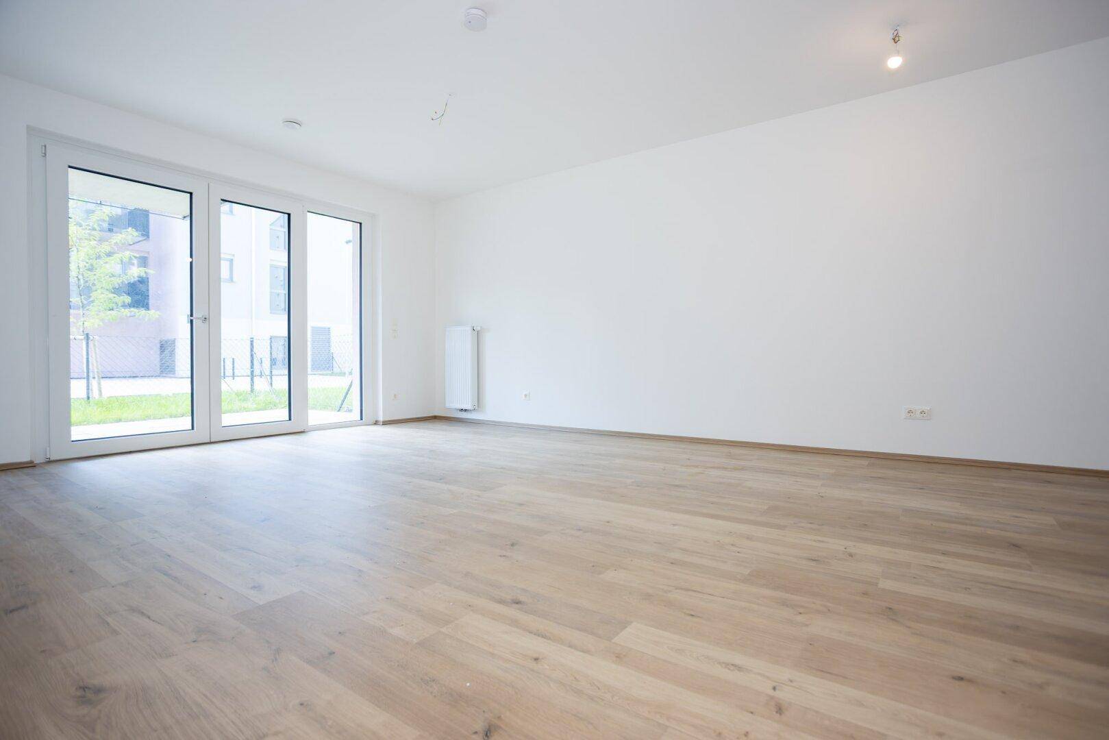 Heller, leerer Wohnraum mit Laminatboden und großer Glastür zum Außenbereich.