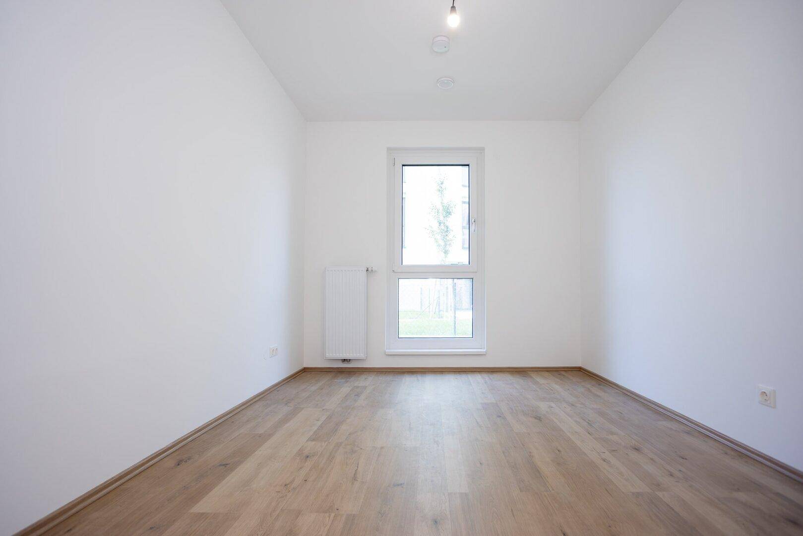 Leeres Schlafzimmer mit hellem Laminatboden und einem Fenster mit Blick ins Grüne.