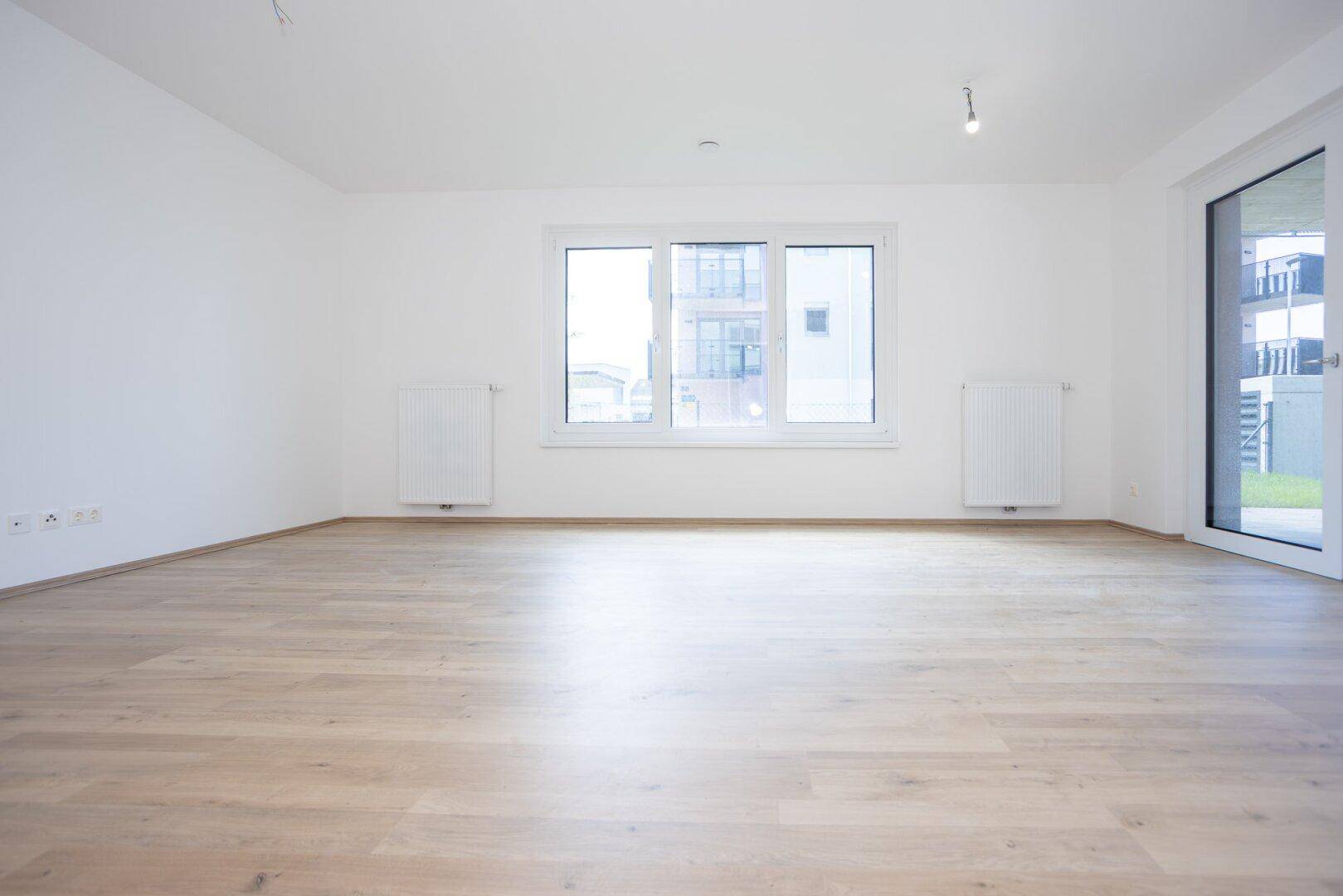 Heller, leerer Wohnraum mit Laminatboden und großen Fenstern in einem Neubau.