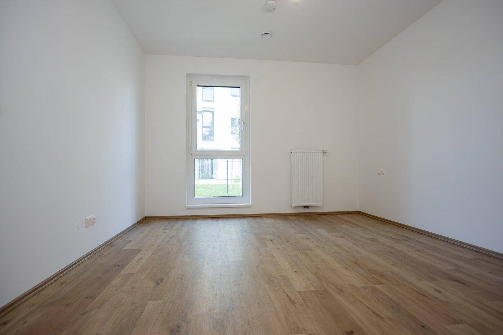 Helles, leeres Zimmer mit Laminatboden und einem Fenster mit Blick auf den Außenbereich.