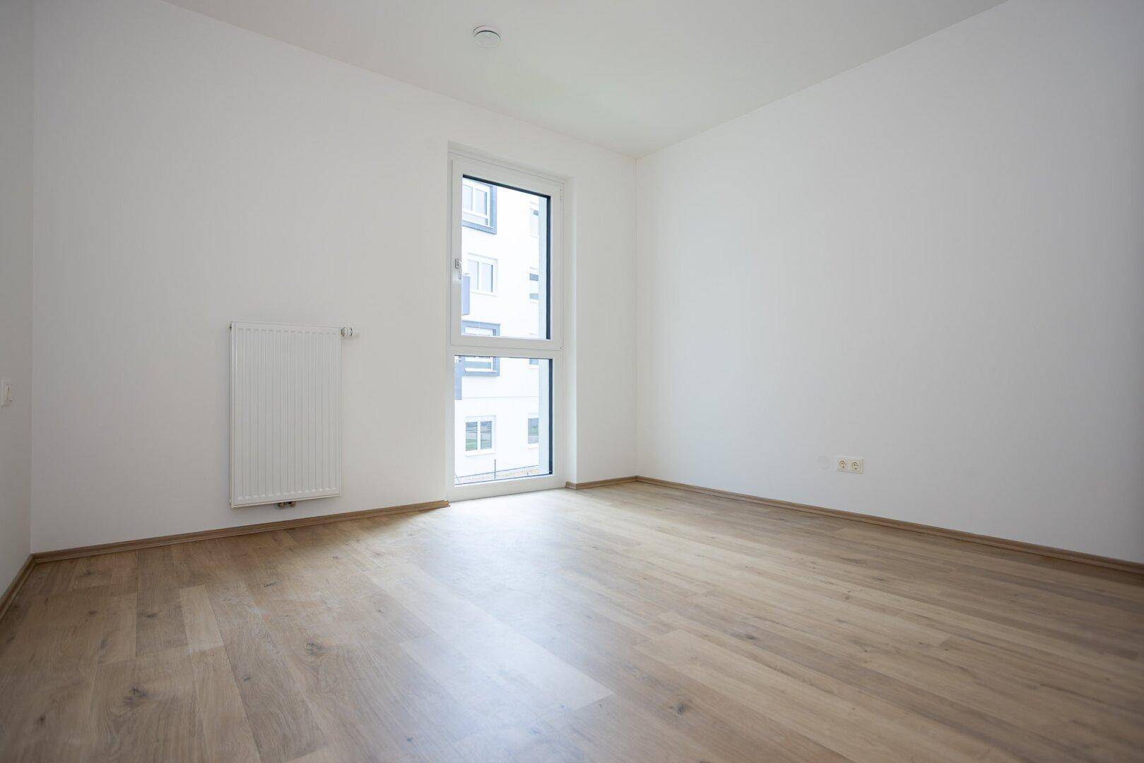 Leeres Zimmer mit hellem Laminatboden und einem Fenster, das viel Tageslicht hereinlässt.