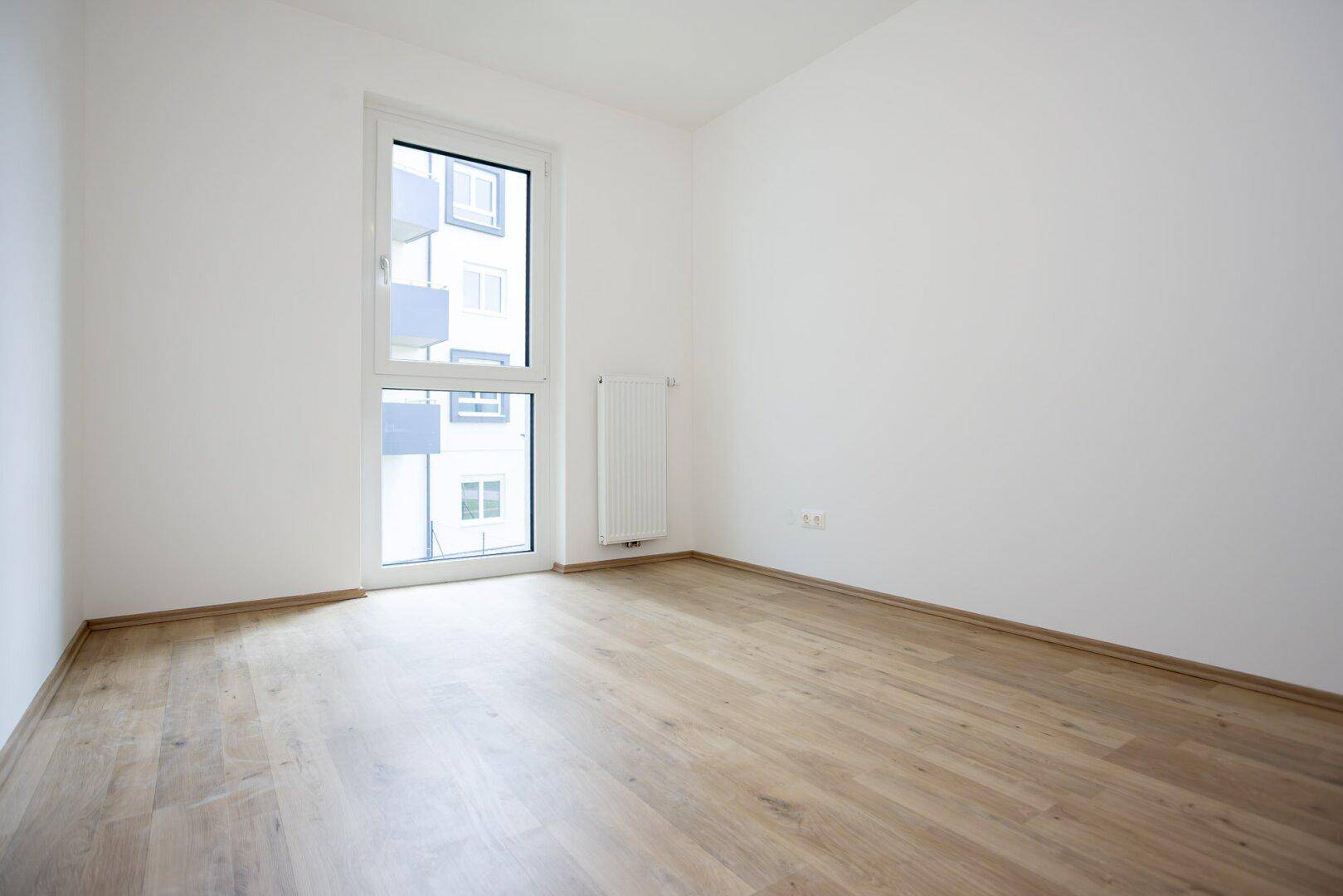 Helles, leeres Zimmer mit Laminatboden und einem hohen Fenster, ideal als Schlaf- oder Arbeitszimmer.