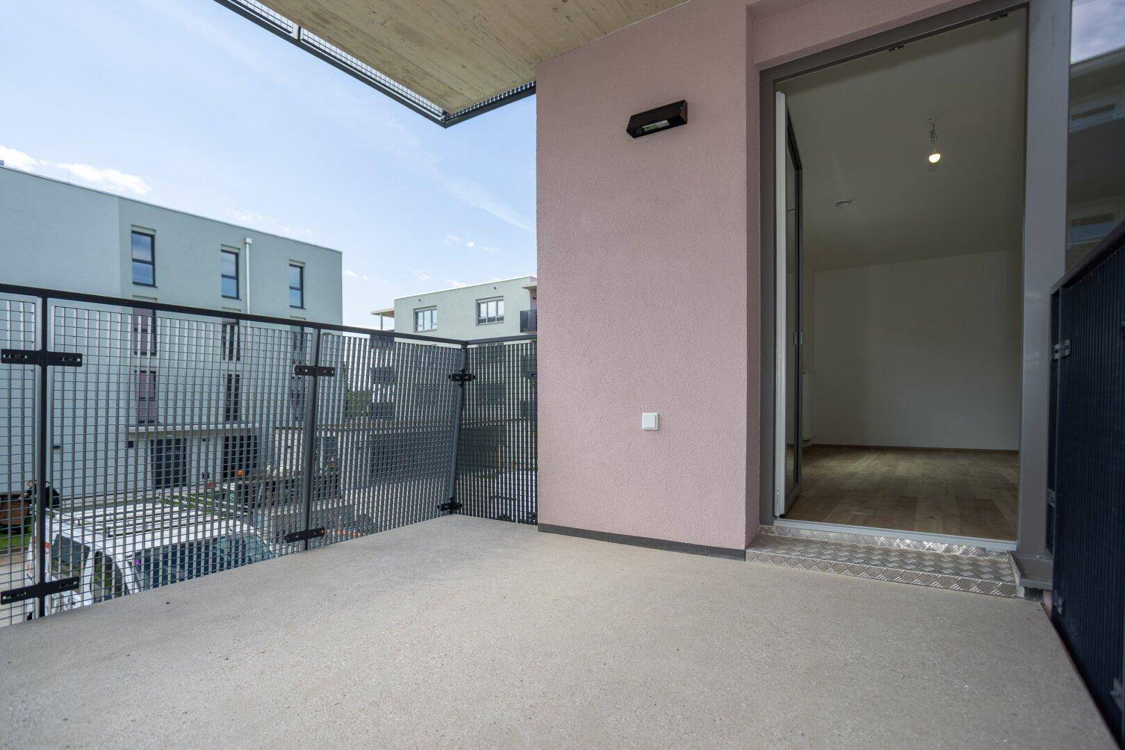 Balkon mit Gittergeländer und Zugang zu einem Innenraum, umgeben von modernen Gebäuden.