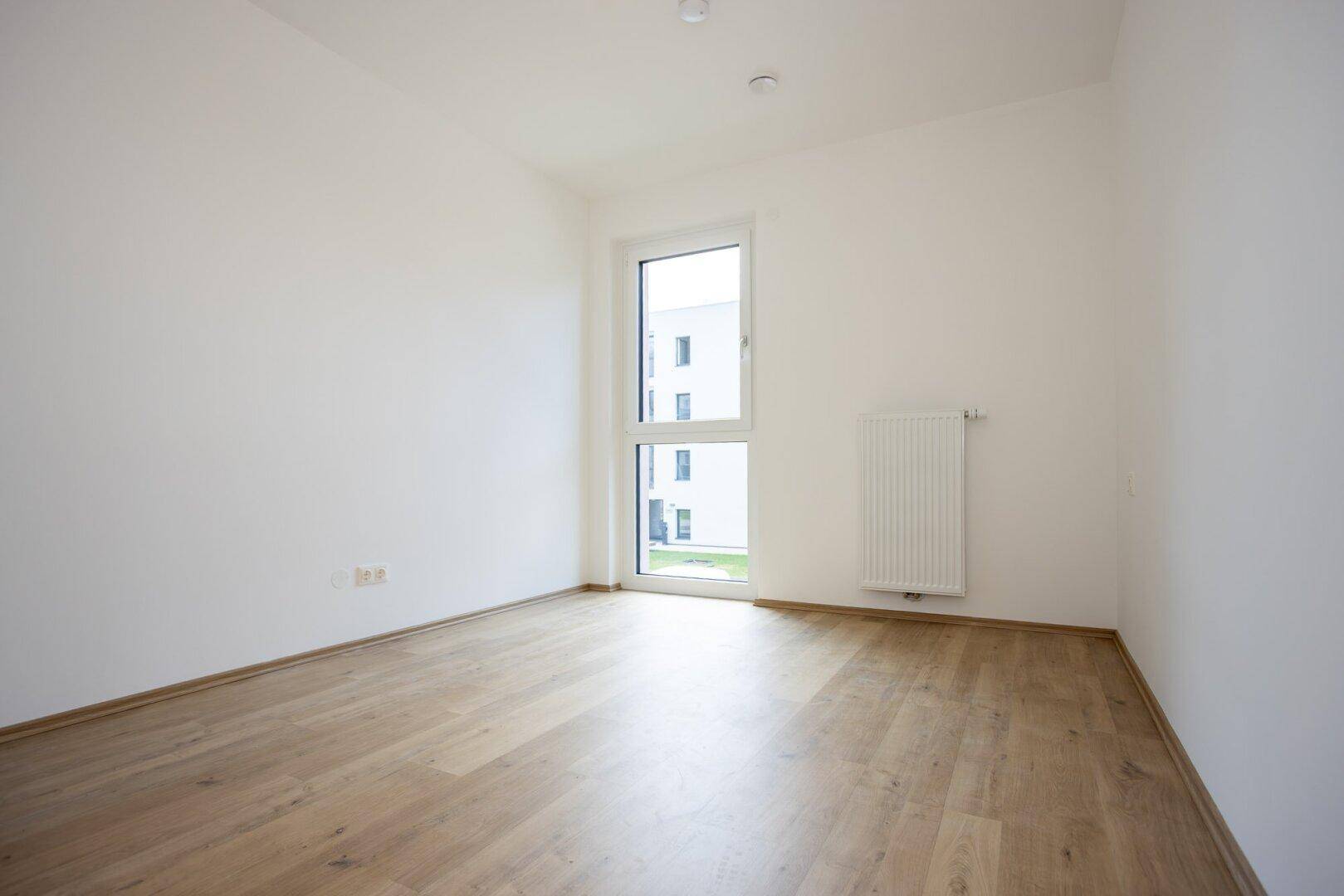 Leeres Zimmer mit hellem Laminatboden, weißen Wänden und einem hohen Fenster für viel Tageslicht.