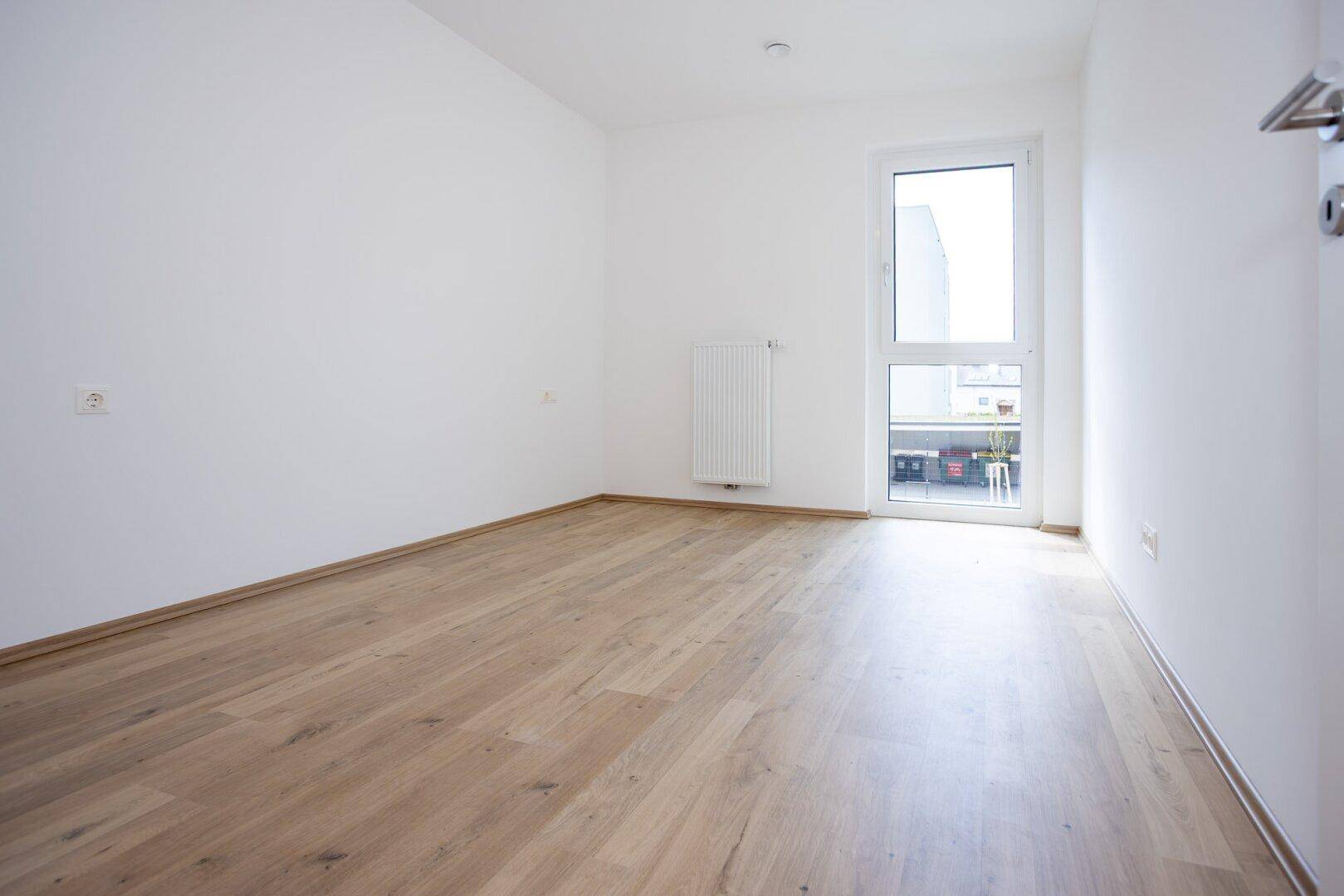 Helles Zimmer mit Laminatboden und bodentiefem Fenster, das viel Tageslicht hereinlässt.